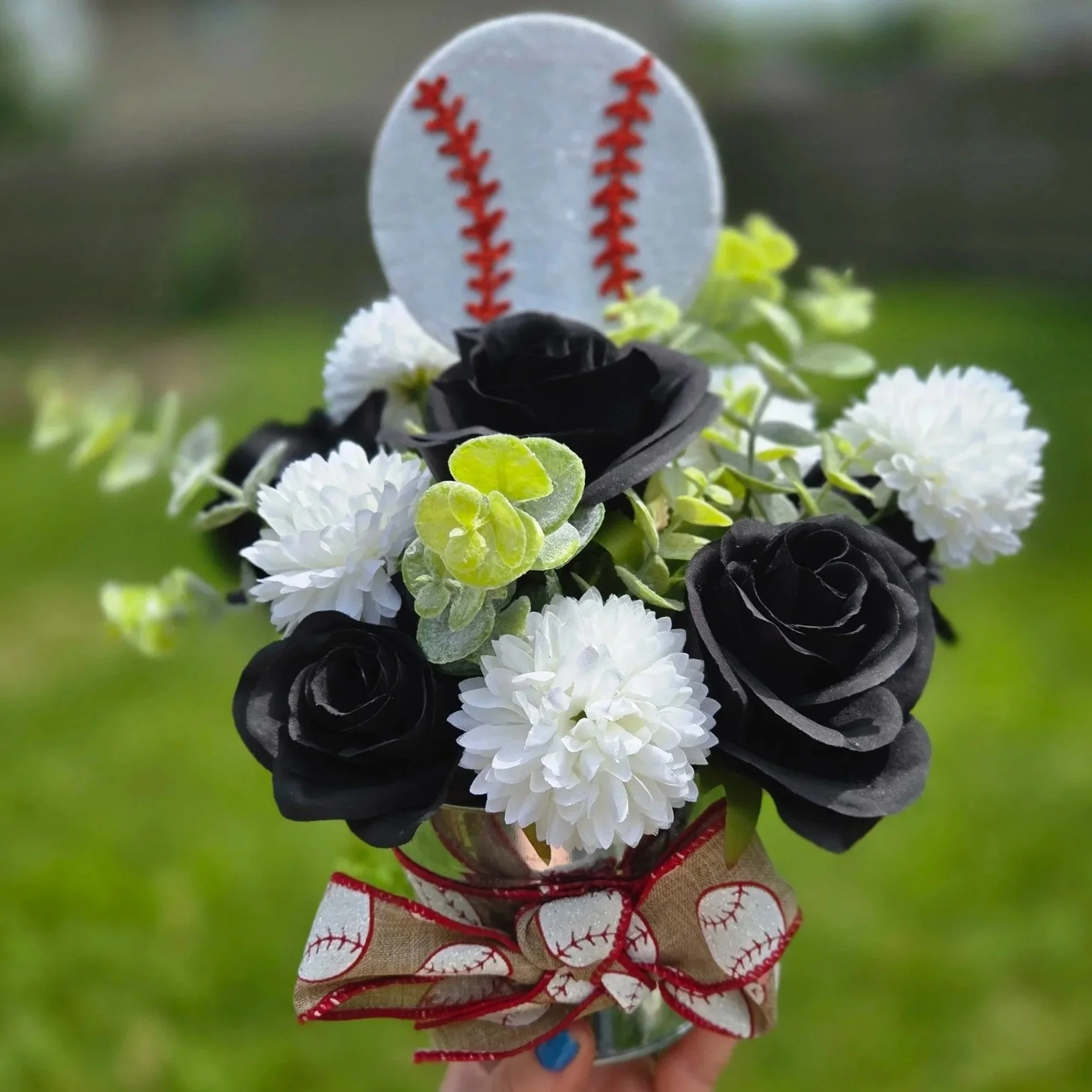 A baseball-themed figurine with a baseball cap, surrounded by white and black flowers with green leaves, held in a person's hand with painted blue nails.