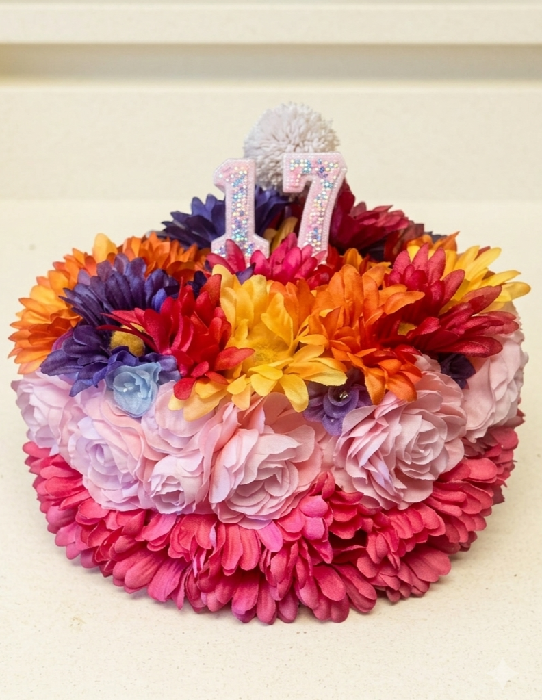 A colorful flower arrangement in a round shape with pink, purple, orange, yellow, and red flowers surrounding a number 17 decoration with a white pom-pom on top, celebrating a 17th birthday.