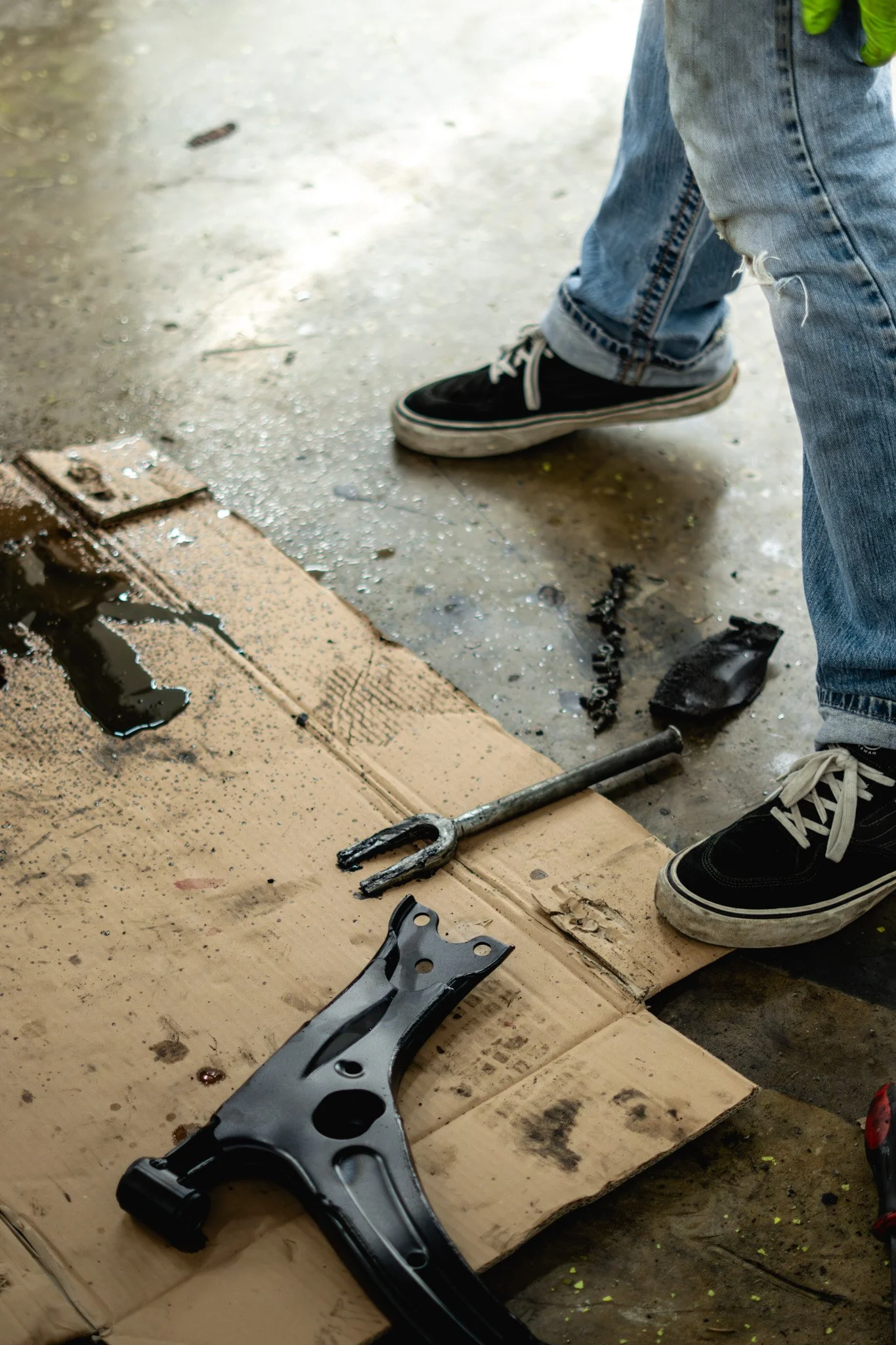 Tools and parts on a cardboard surface, a person wearing blue jeans and black sneakers standing nearby, with some liquid and debris on the ground.