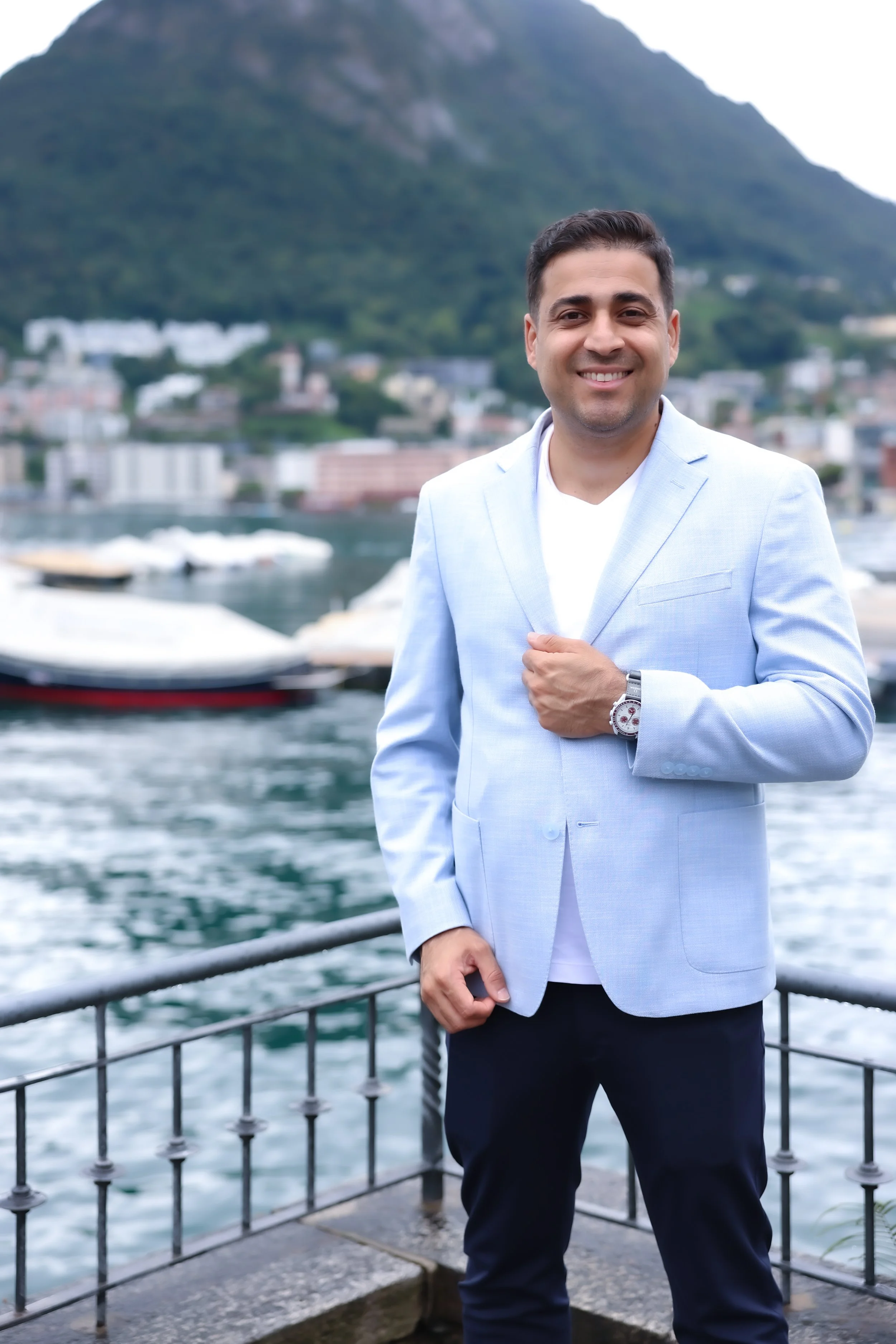 A man in a light blue blazer and dark pants standing on a boat dock with a body of water and boats in the background, smiling at the camera.