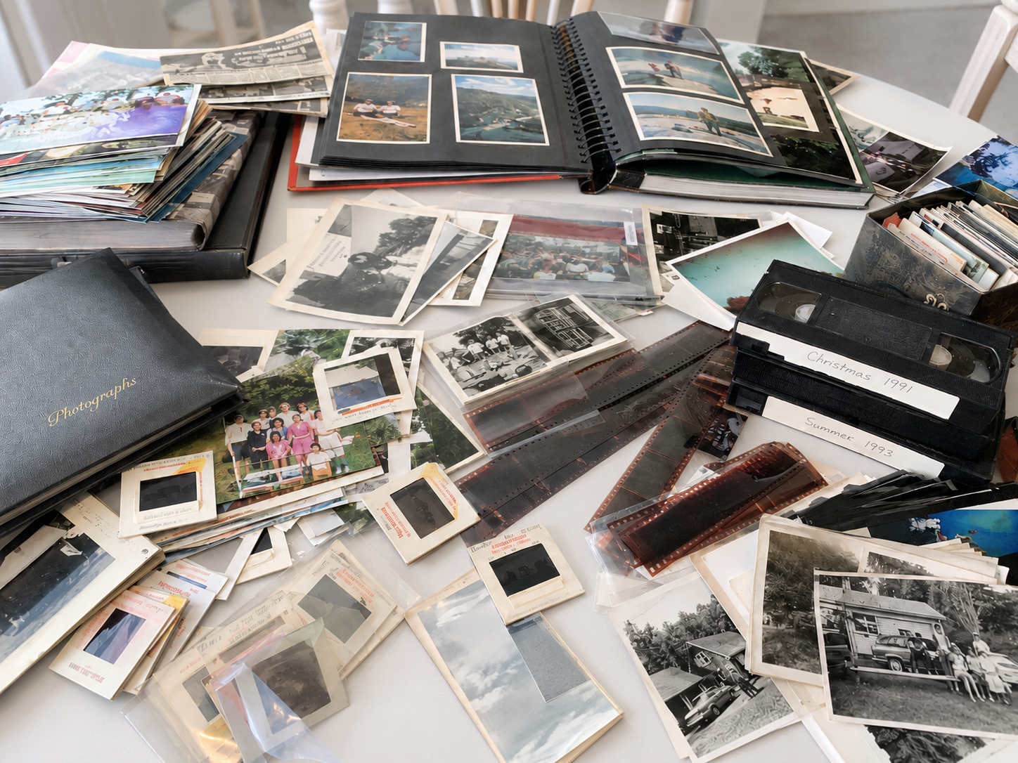 A table filled with numerous photo albums, loose photographs, and film slides, some labeled with dates like 'Christmas 1991' and 'Summer 1993', depicting family and outdoor scenes. | My Memory File | Cheryl DiFrank | Professional Photo Manager
