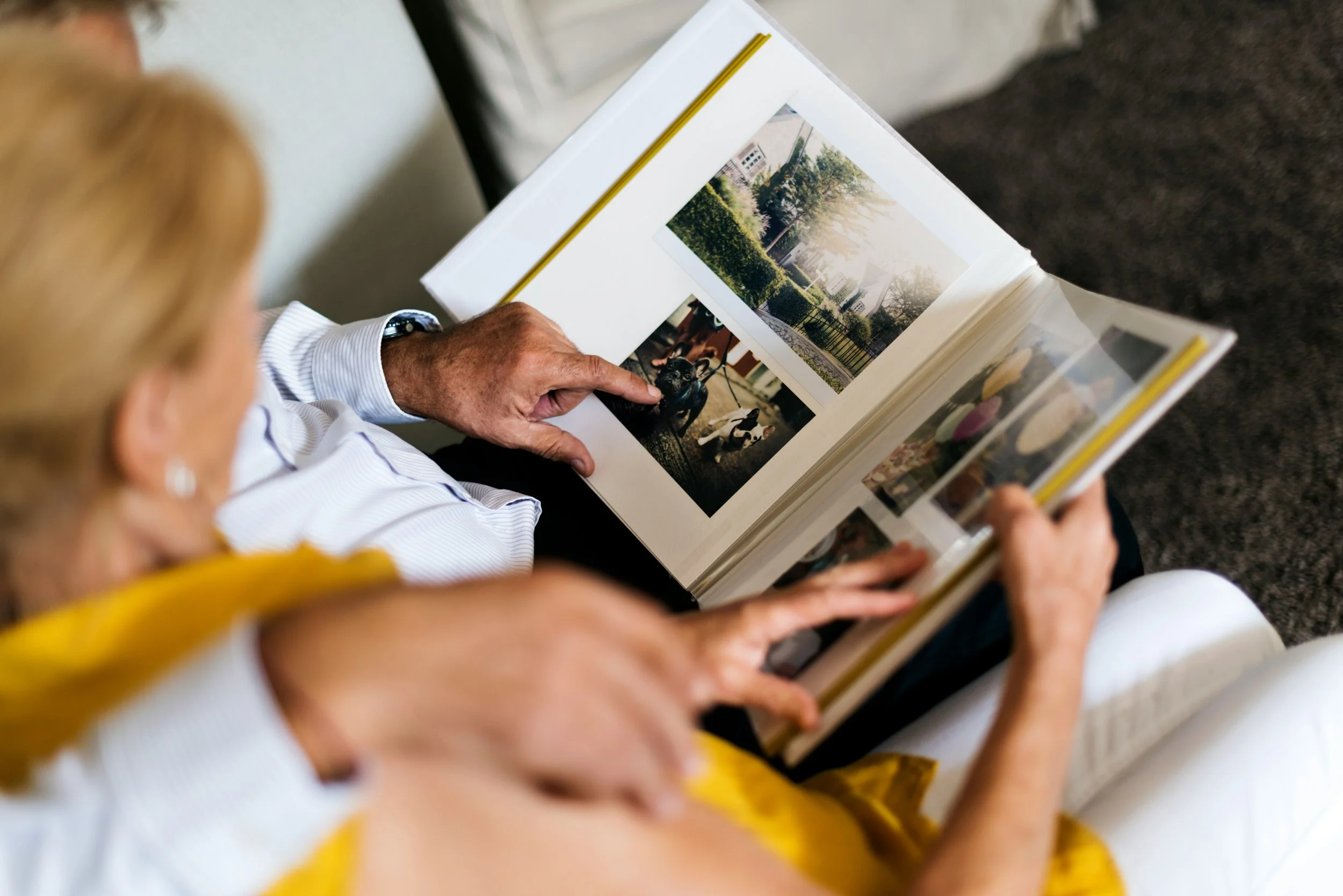 Close-up of an elderly woman and a person with blonde hair looking at a photo album together. The photo album contains images of a park with trees and a dog.