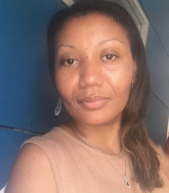 A woman with long hair, wearing earrings, a necklace, and a beige top, standing in front of a blue background.