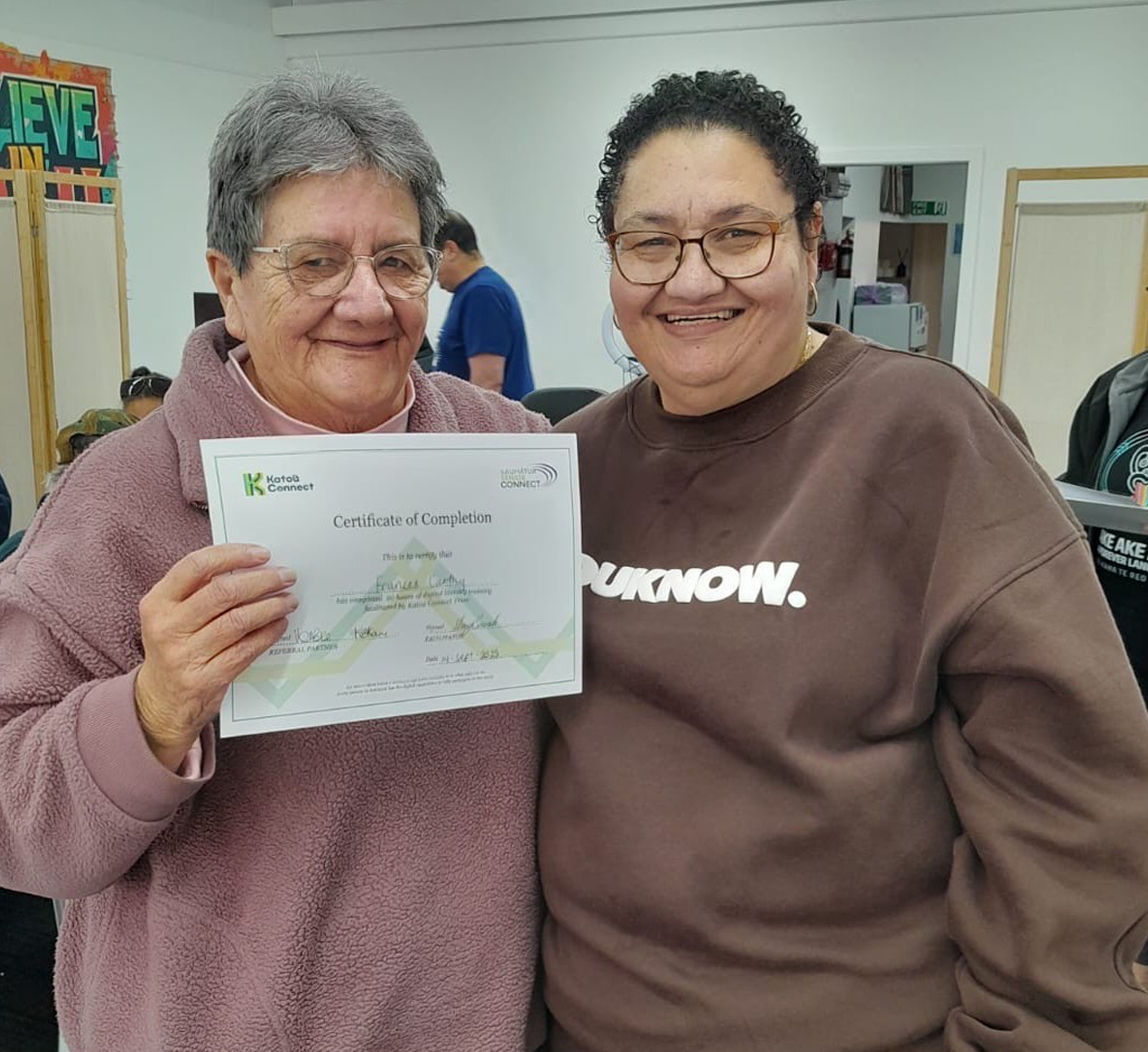 Two smiling people stand together indoors, with one proudly holding a Katoa Connect certificate of completion. The image celebrates learning, achievement, and community support.
