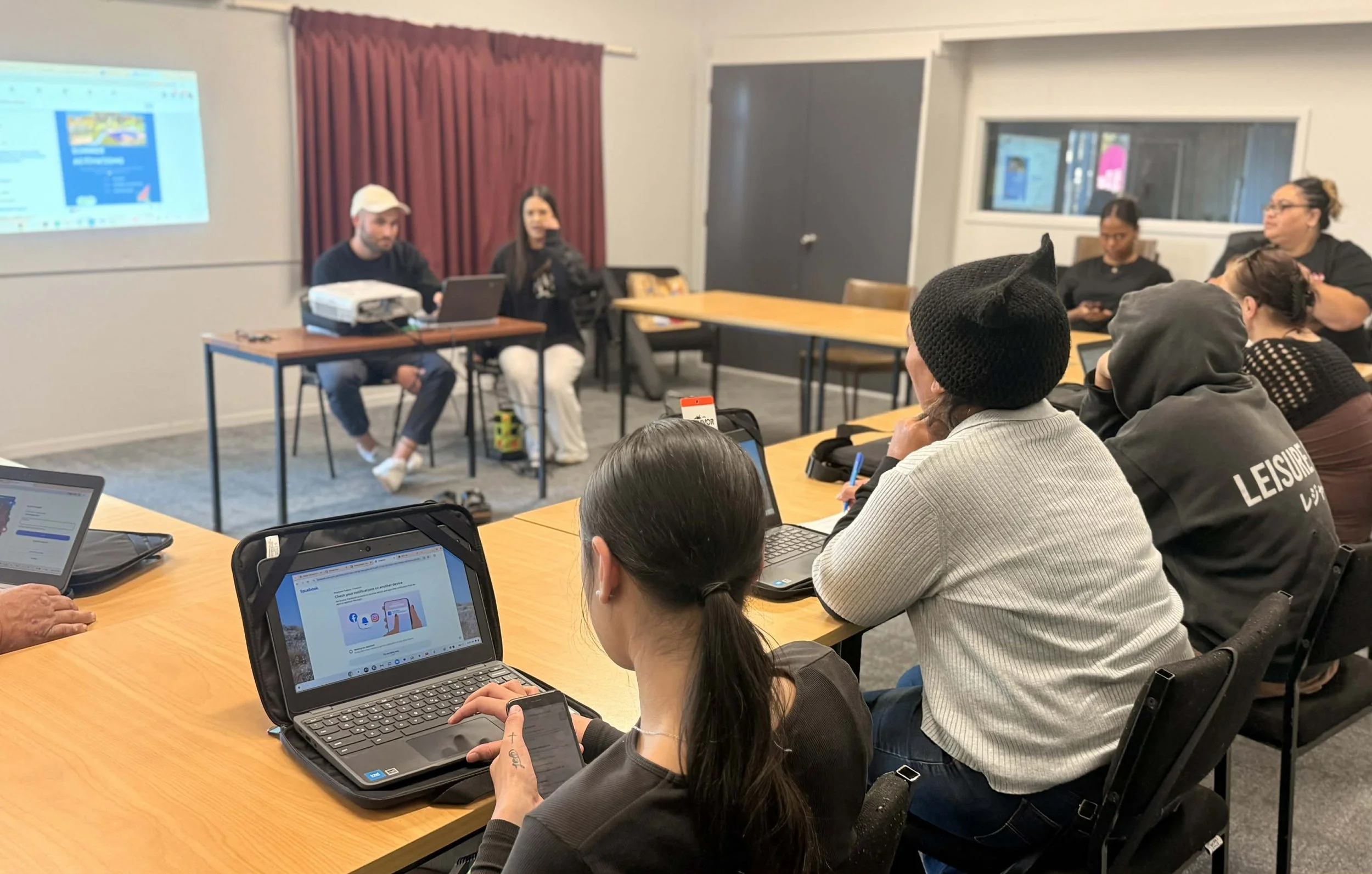 People participate in a Katoa Connect digital skills workshop, using laptops and devices while following a presentation at the front of the room. The image highlights hands-on learning, digital inclusion, and community education.