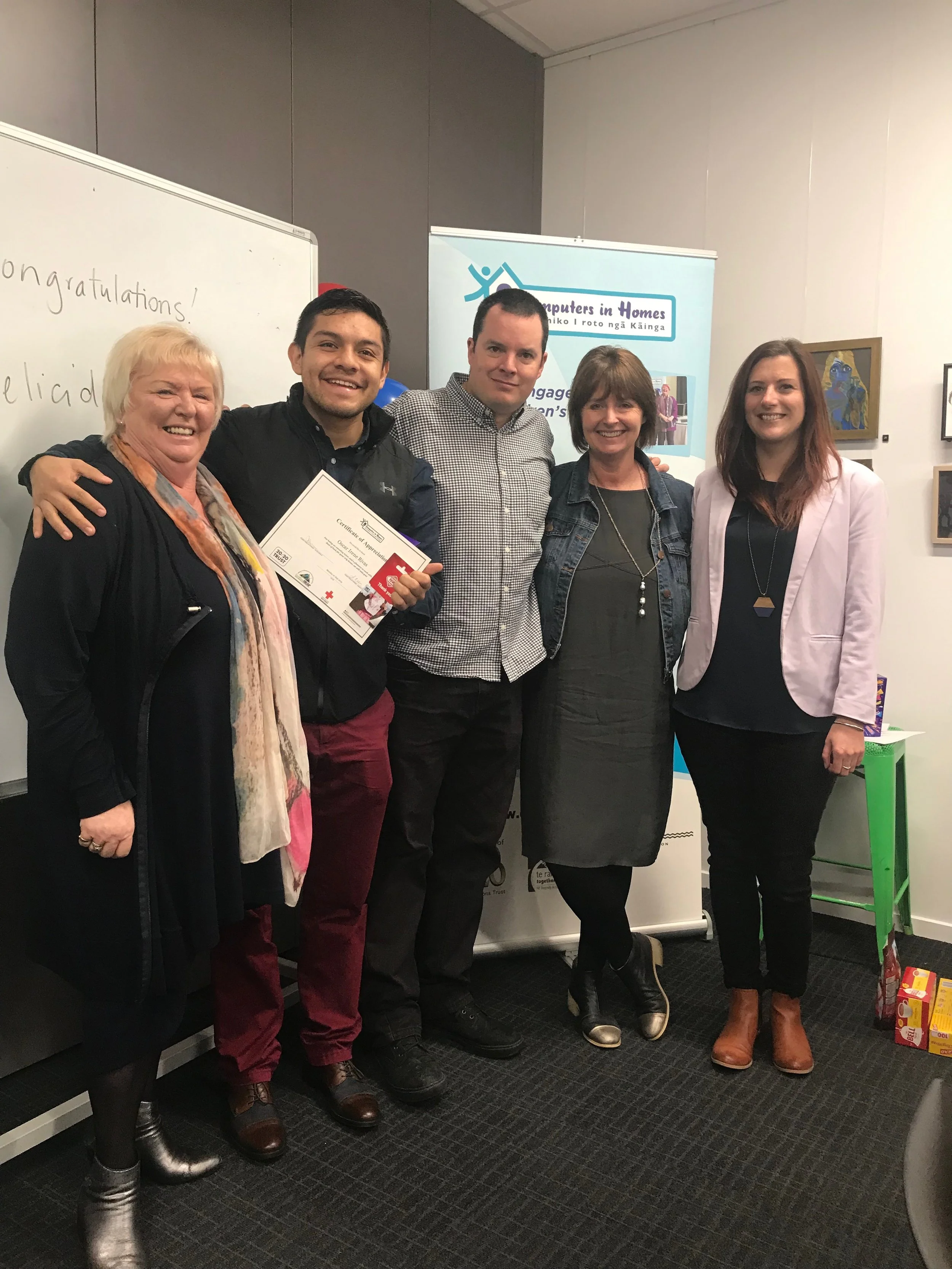 A group of five smiling people standing together indoors, with some holding a certificate, in front of a whiteboard and a banner.
