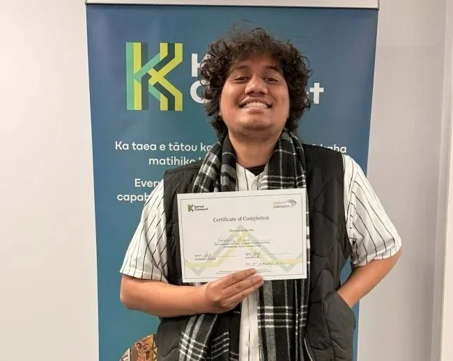 A smiling person stands proudly in front of a Katoa Connect banner, holding a certificate of completion. They are wearing a striped shirt, scarf, and black vest, celebrating their achievement.
