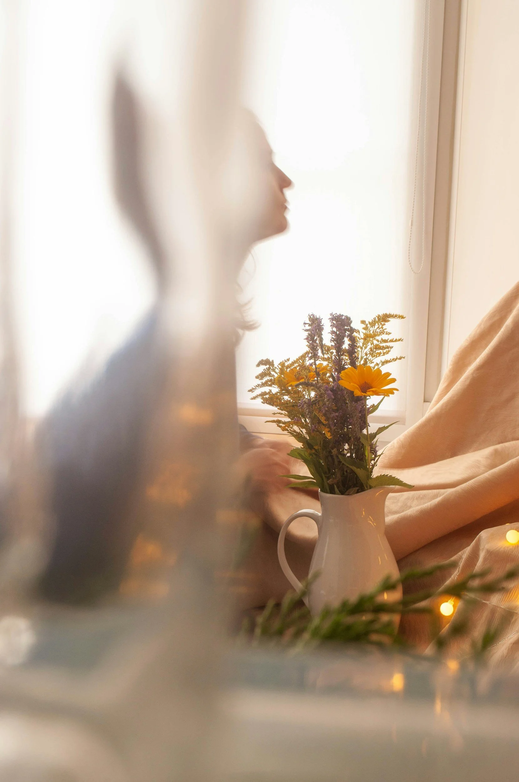 A woman with long hair in profile sits near a window with sheer curtains, holding a bouquet of purple and yellow flowers in a white pitcher. Soft, warm lighting and string lights create a cozy atmosphere.