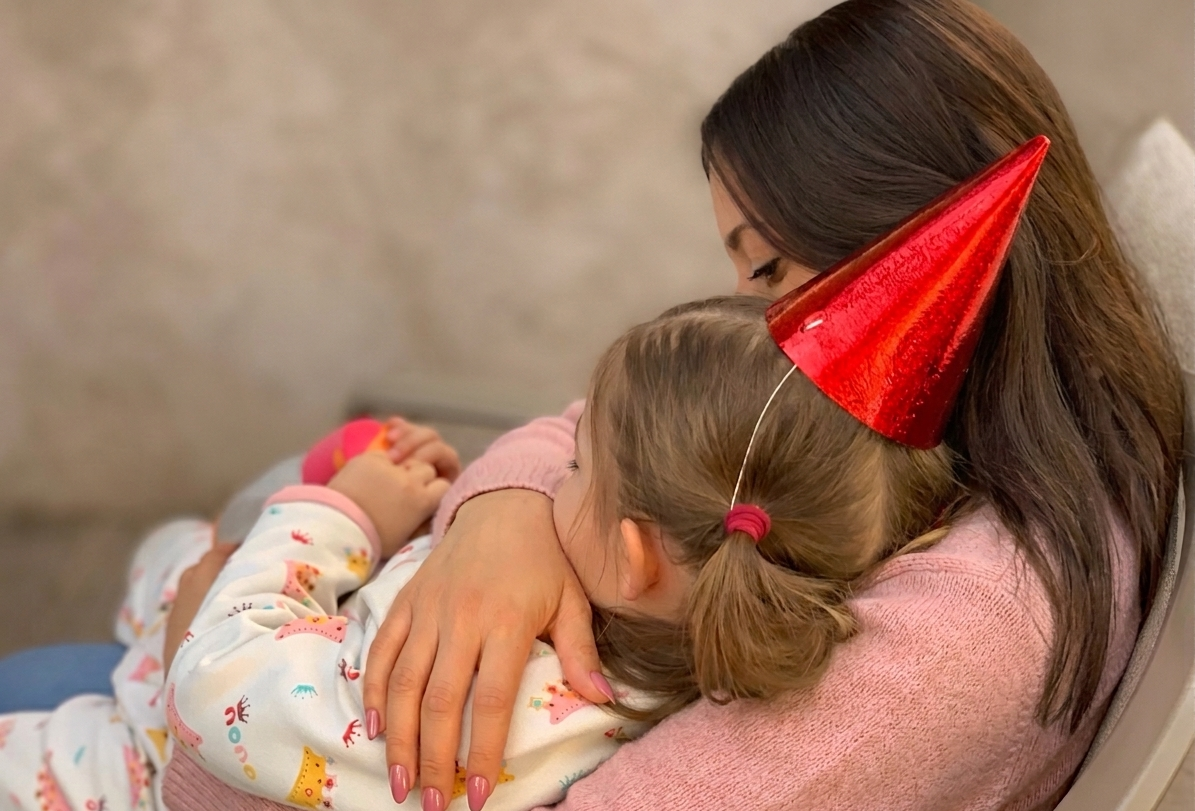 A woman with long brown hair wearing a red party hat is hugging a young girl with light brown hair tied in a ponytail. The girl is wearing a party hat and pajamas with colorful patterns. They are sitting on a chair or couch, embracing in a warm hug.