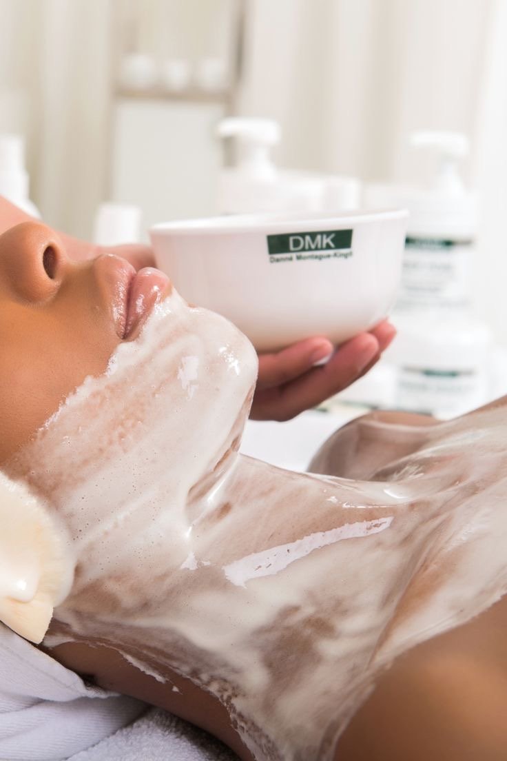 Person receiving a facial treatment with foam or cleanser, lying down with a bowl labeled 'DMK' in the background.