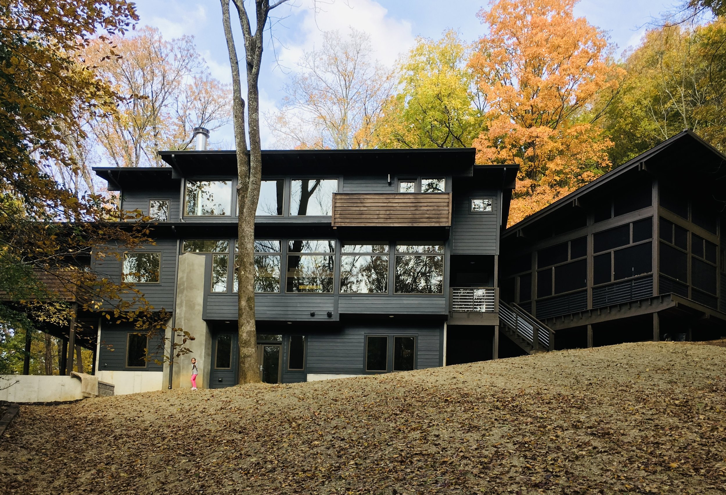 Burke Architecture & Design Associates modern custom residential home design design featuring large windows and gray siding is situated on a hillside, surrounded by trees with autumn-colored leaves.