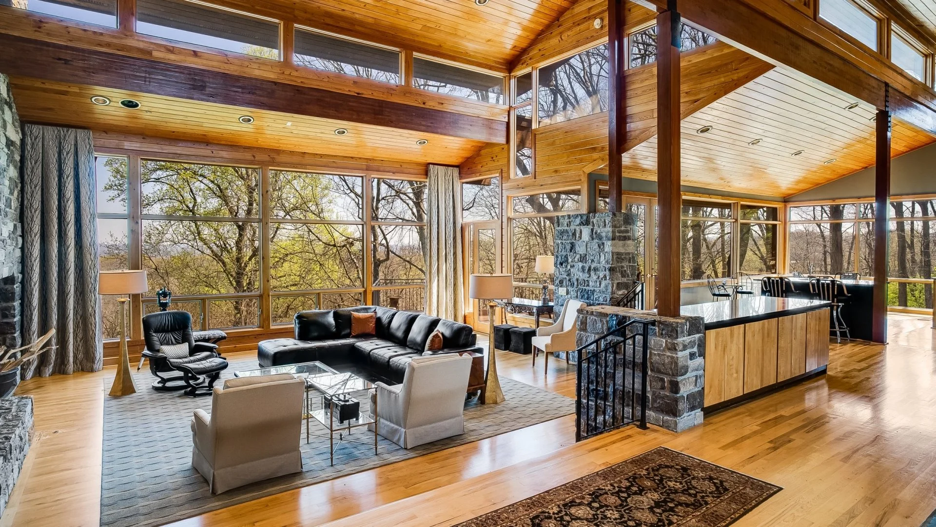 Custom home design across Middle TN by Burke Architecture & Design Associates featuring vast windows, wood-beamed ceilings, wood floors, stone fireplace, black leather sofa, white armchairs, and a large kitchen island with barstools.