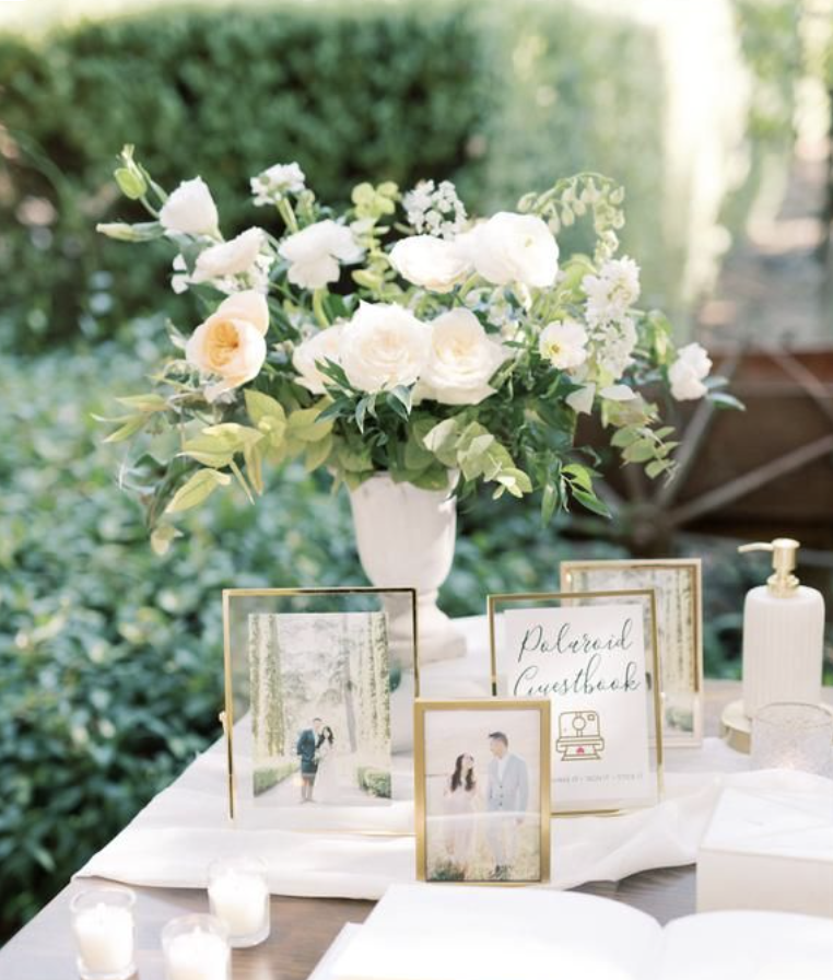 Table with a large floral arrangement of white flowers and green leaves, framed photographs of a couple, a sign that says "Polaroid Guestbook," candles, and a white tablecloth outdoors in a garden setting.