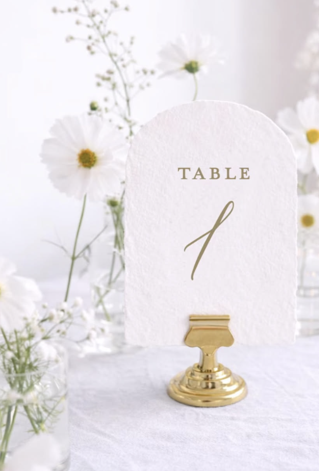 Table number card with a gold stand, decorated with white flowers in the background.