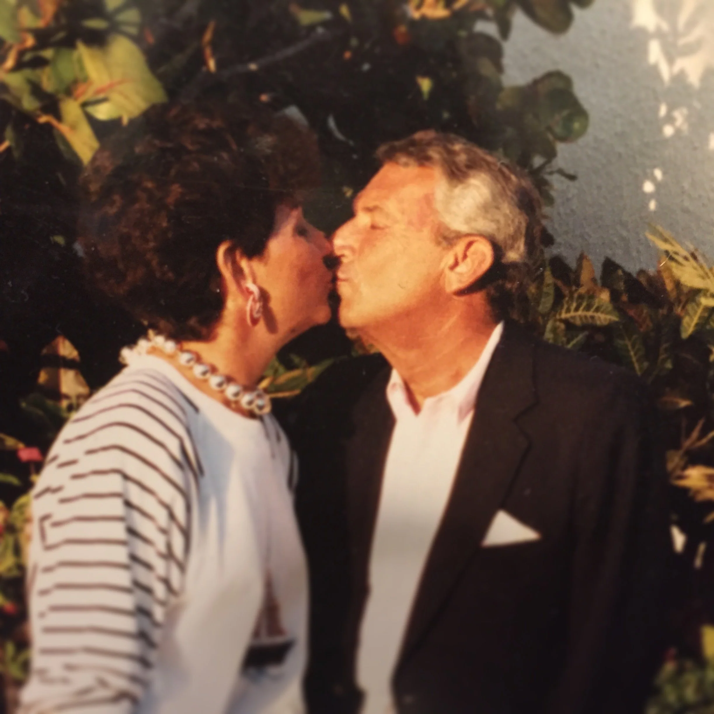 A couple is about to kiss outdoors, surrounded by leaves, with their faces close together, eyes closed, and noses almost touching.