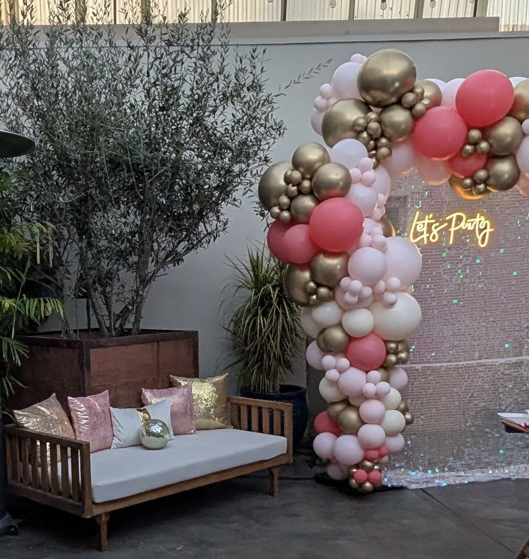 Decorative balloon arch with pink, white, and gold balloons next to a seating area with pillows and a neon sign that reads 'Let's Party'.