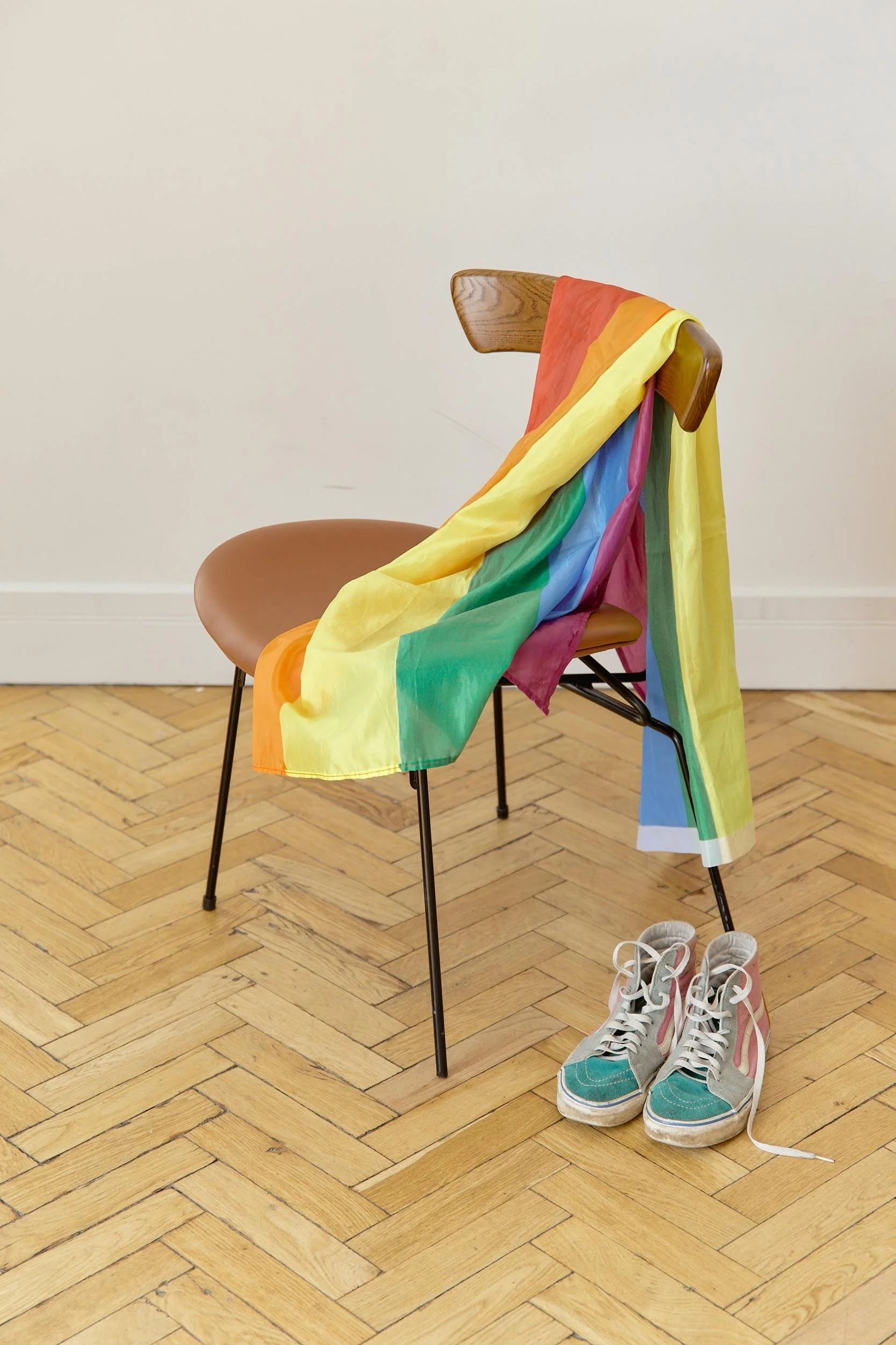 A wooden chair with a rainbow flag draped over it, a pair of sneakers on the floor beside it, and a pair of shoes in the background.