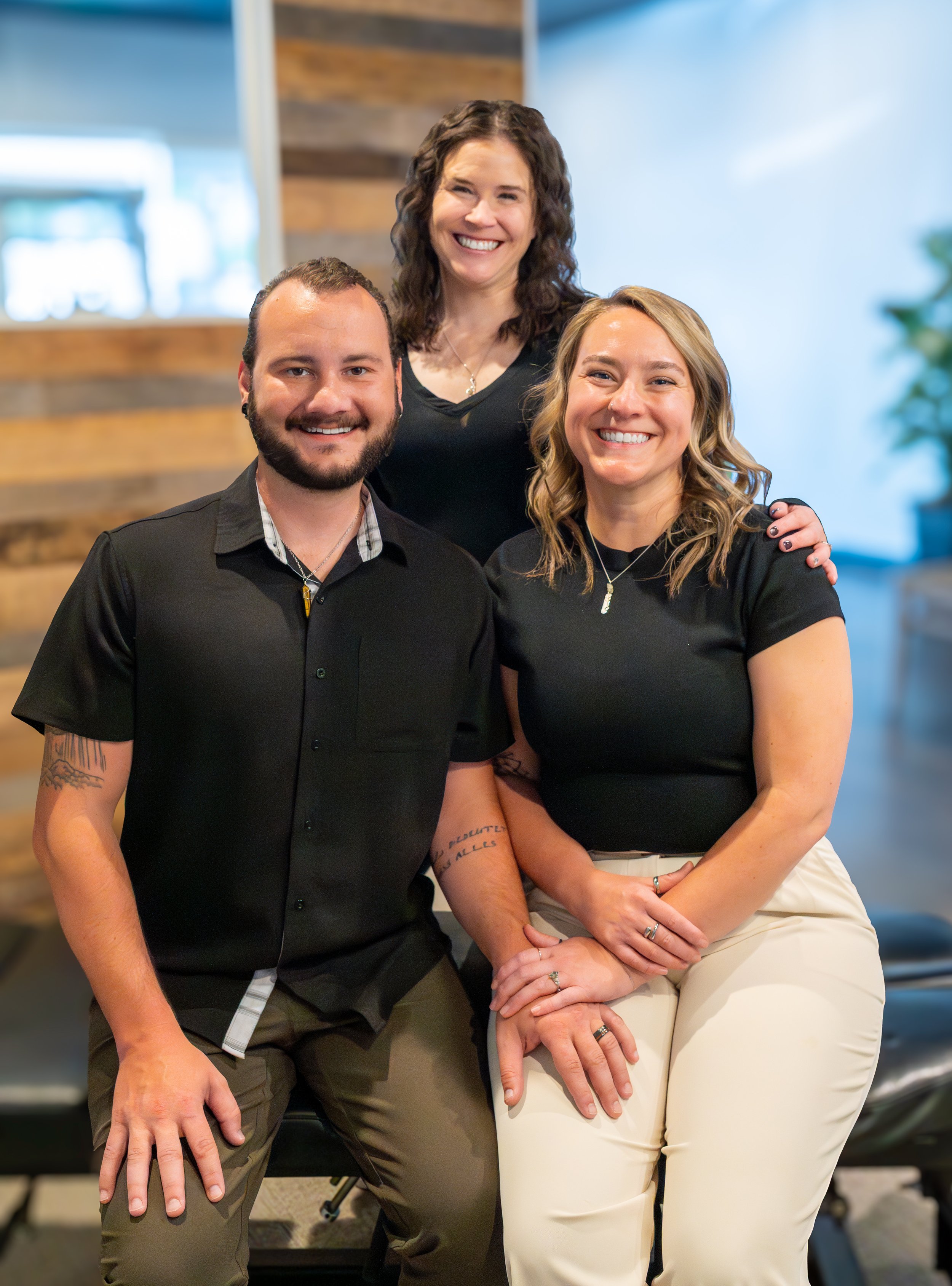 Three smiling people, two women and one man, posing together indoors. The man is wearing a black shirt, the woman on the right is wearing a black top and beige pants, and the woman in the middle is wearing a black top. They are close together, with the man and woman on the right sitting and the woman in the middle standing behind them with her arms on their shoulders.