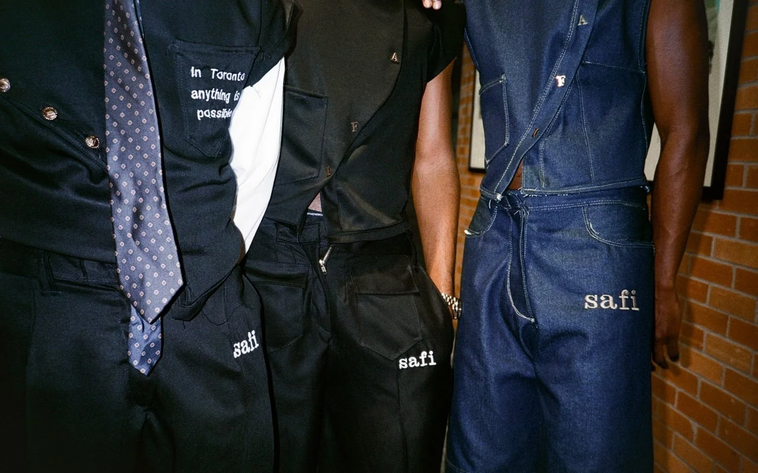 Close-up of three people wearing denim and black clothing, with the word 'safi' embroidered on their pants and denim vest, standing against a brick wall.