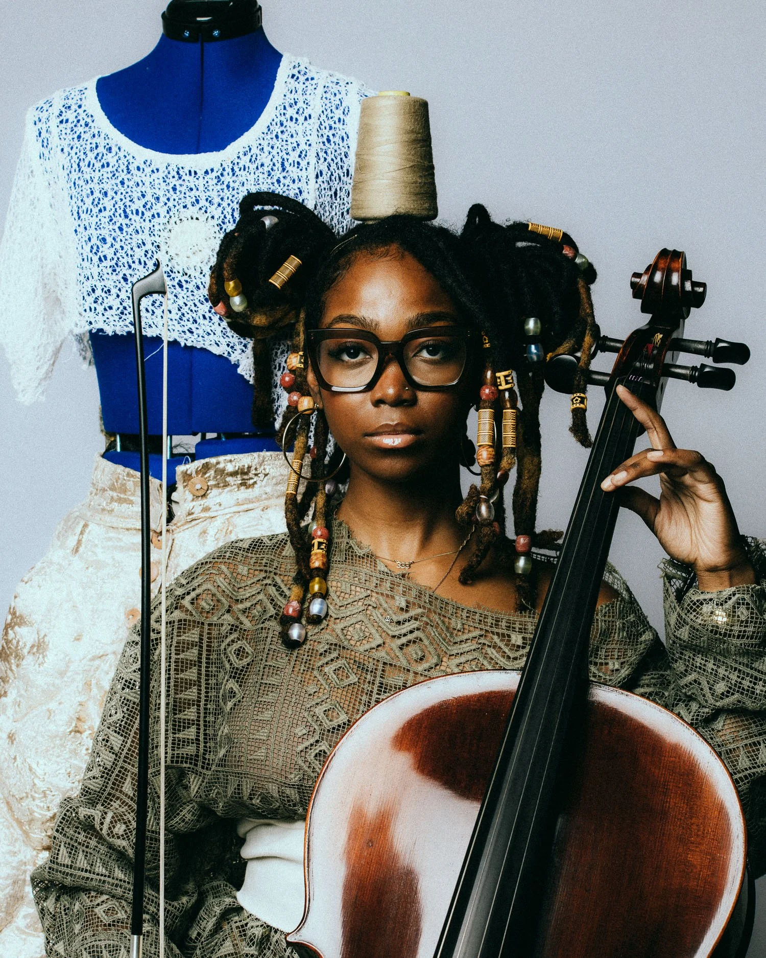 A woman with dreadlocks holding a cello, wearing glasses and a patterned top, standing in front of a mannequin dressed in a white lace top.