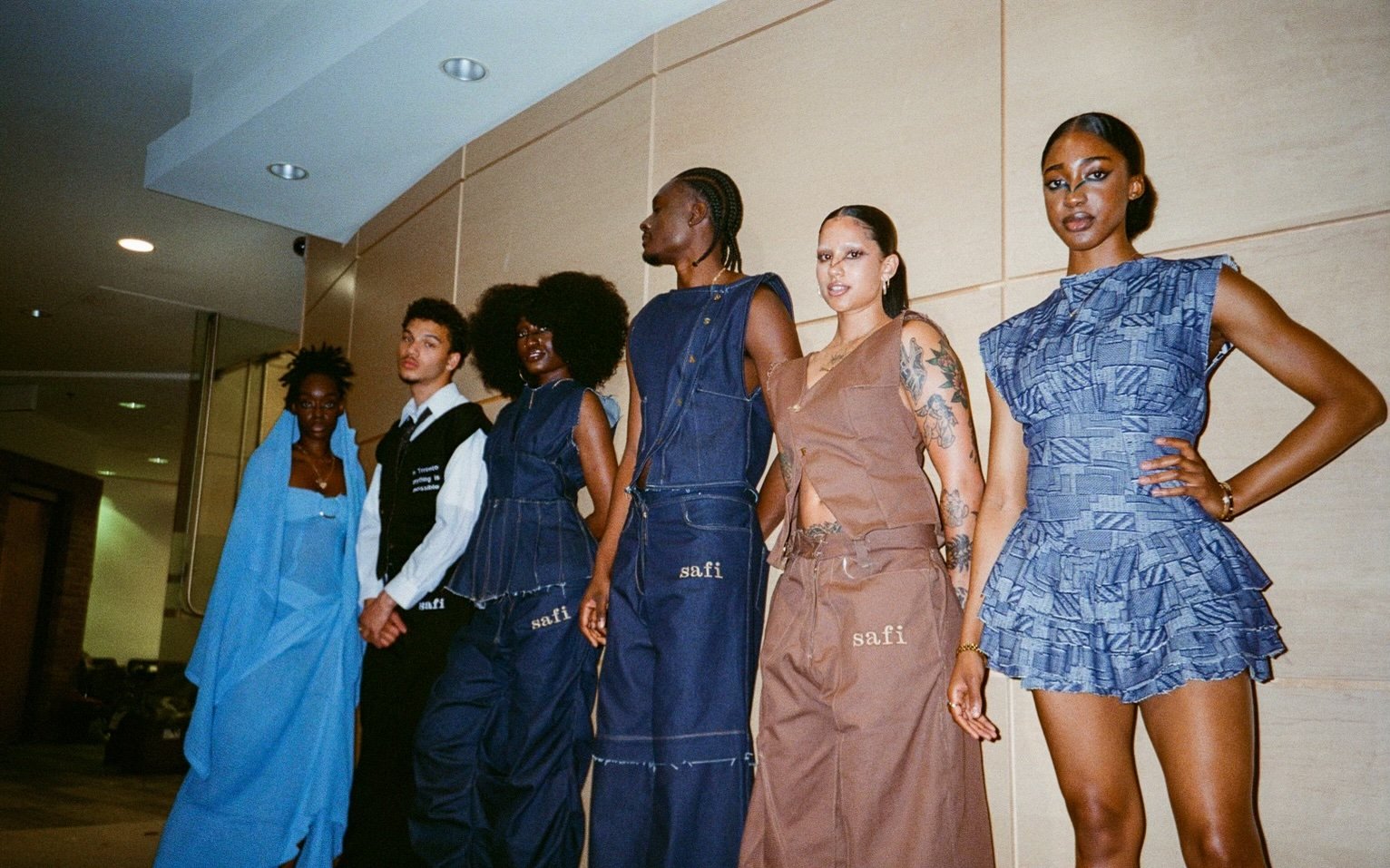 Six diverse models standing in a line indoors, showcasing fashion designs, with varied poses and expressions.