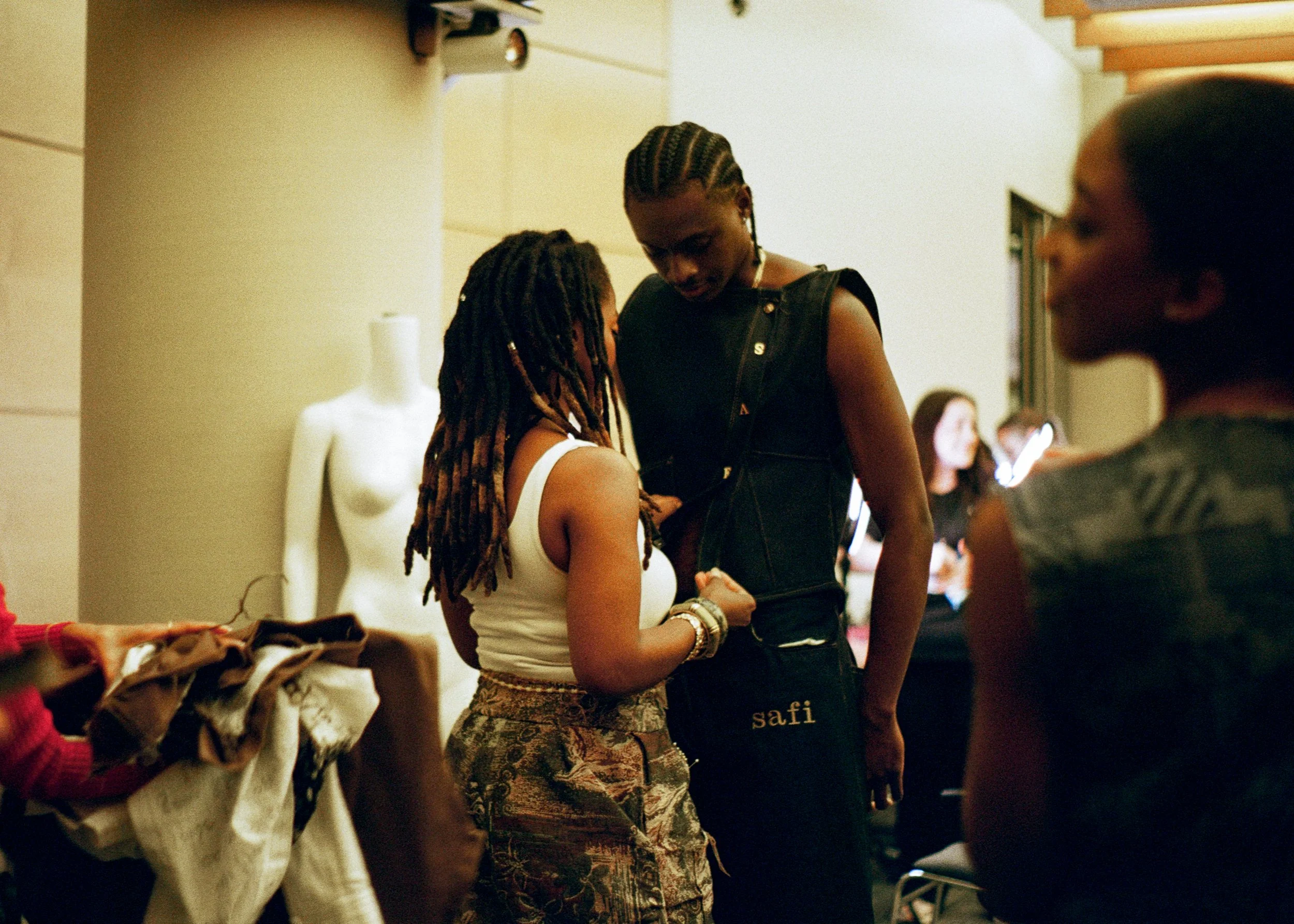 Two women with dreadlocks are standing close together in a store or market, engaging in conversation. One woman is wearing a white tank top and patterned pants, the other is wearing a black vest with gold buttons and the name 'safi' on her pants. There are clothing items on the table in front of them, and mannequins and customers can be seen in the background.