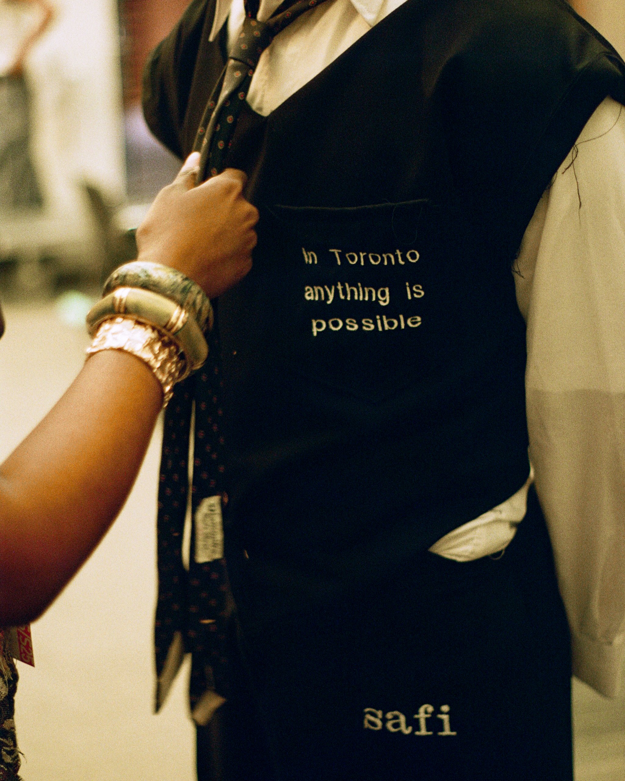 Close-up of a person wearing a black vest with white text that reads "In Toronto anything is possible," and a white shirt underneath, holding the collar of the vest. The person is wearing multiple bracelets on their wrist.