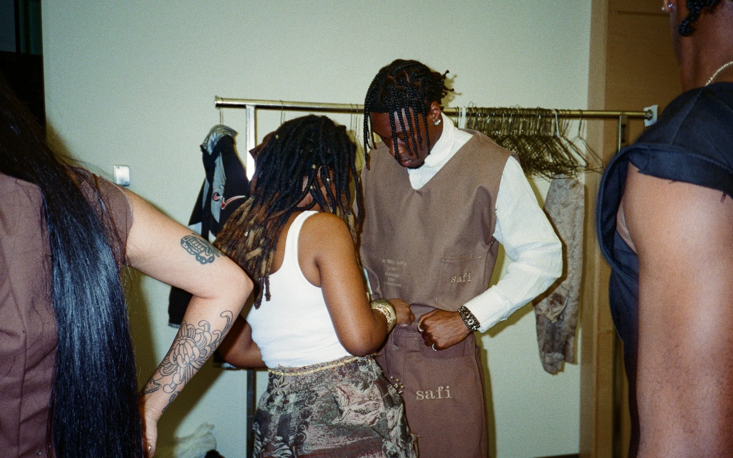 Two people with long dreadlocks sharing an intimate moment, standing in front of a beige wall with a clothing rack behind them.
