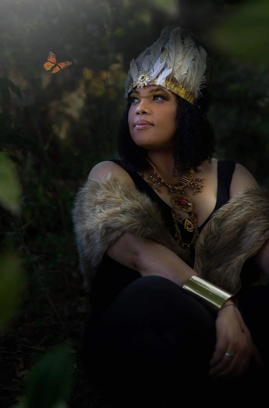 Woman wearing a feathered headdress and jewelry sitting outdoors in a natural setting, looking at a butterfly.
