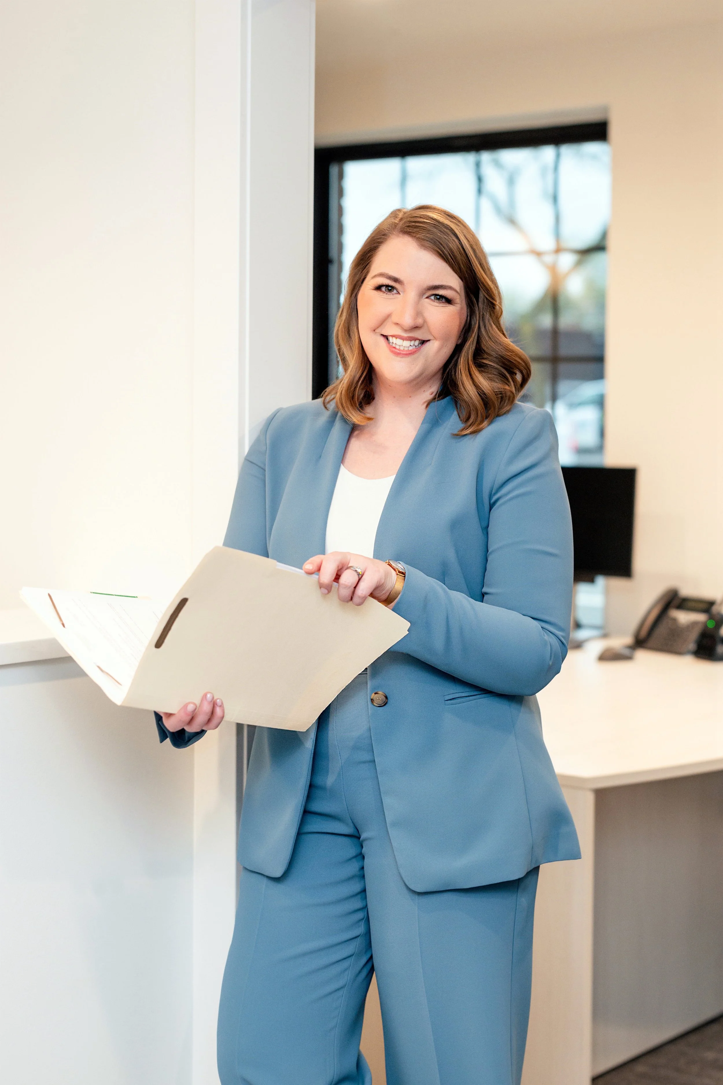 A woman in a blue suit standing in an office, holding a folder and smiling.