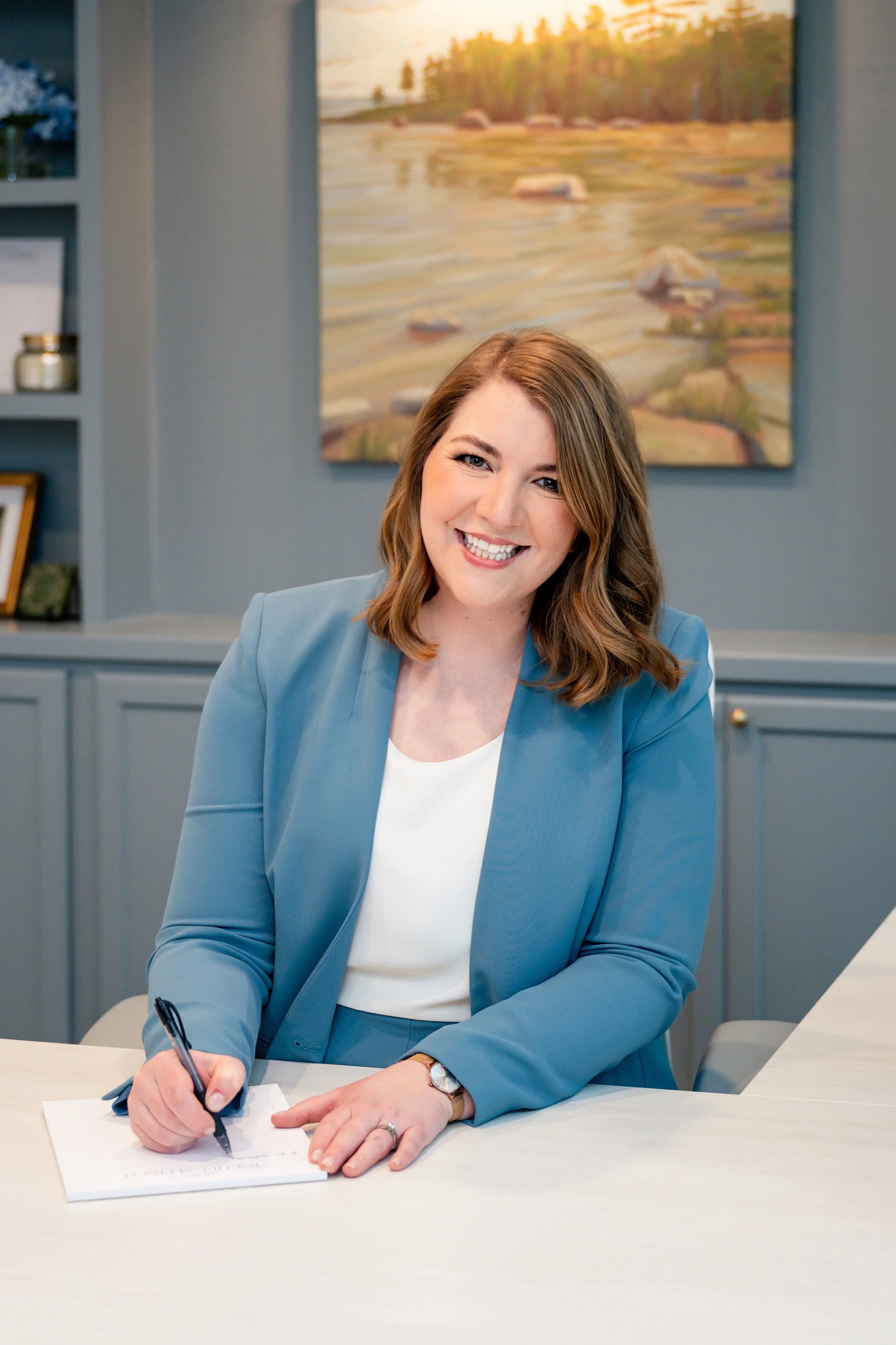 A woman with shoulder-length brown hair smiling while sitting at a white table, wearing a blue blazer and writing on a piece of paper with a pen.