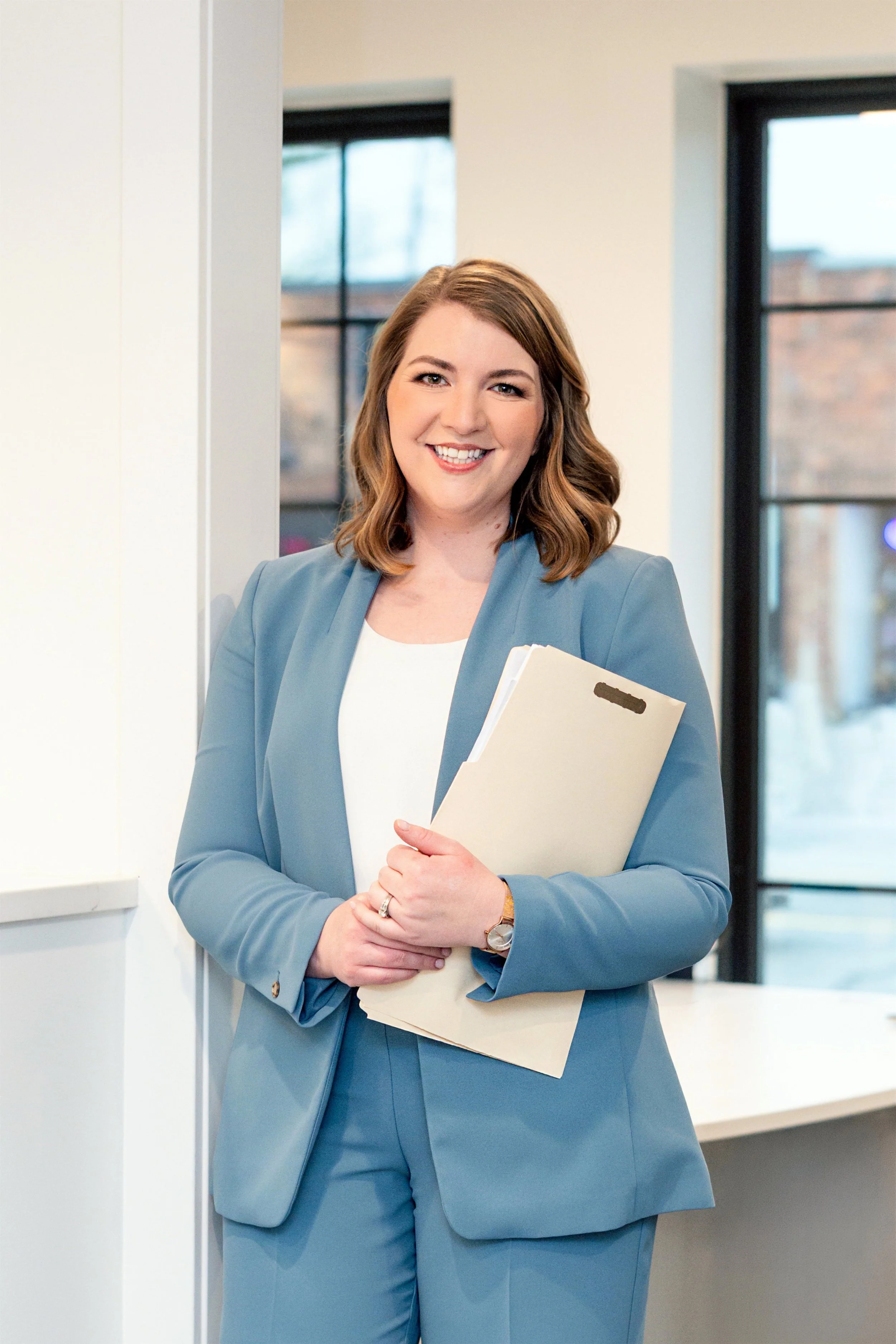 A woman with brown wavy hair smiling, wearing a light blue business suit, holding a folder, standing in front of windows in an indoor setting.