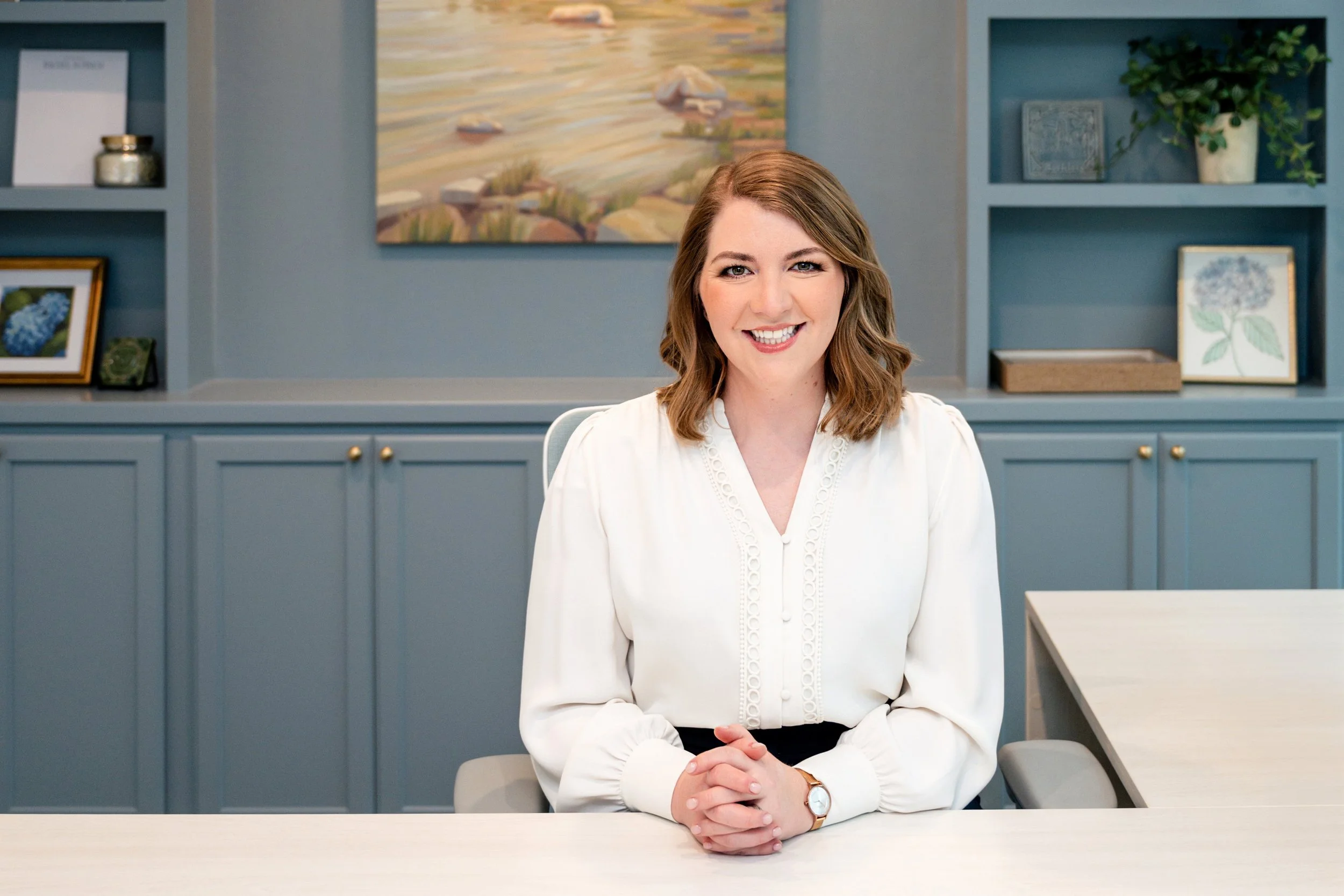 A woman with shoulder-length light brown hair and a white blouse seated at a light-colored desk in an office, smiling at the camera. Behind her is a blue wall with shelves containing framed pictures, a potted plant, and decorative items.