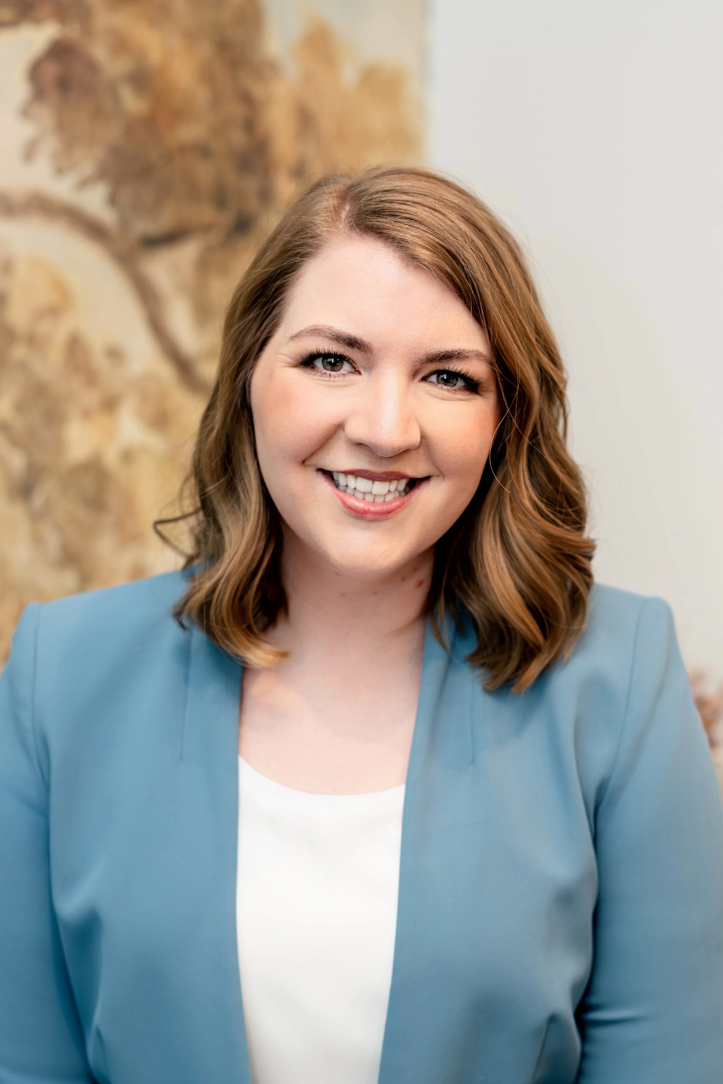 A woman with red hair, blue eyes, and a bright smile wearing a light blue blazer and white top, standing in front of a background with a blurred painting or wall decor.