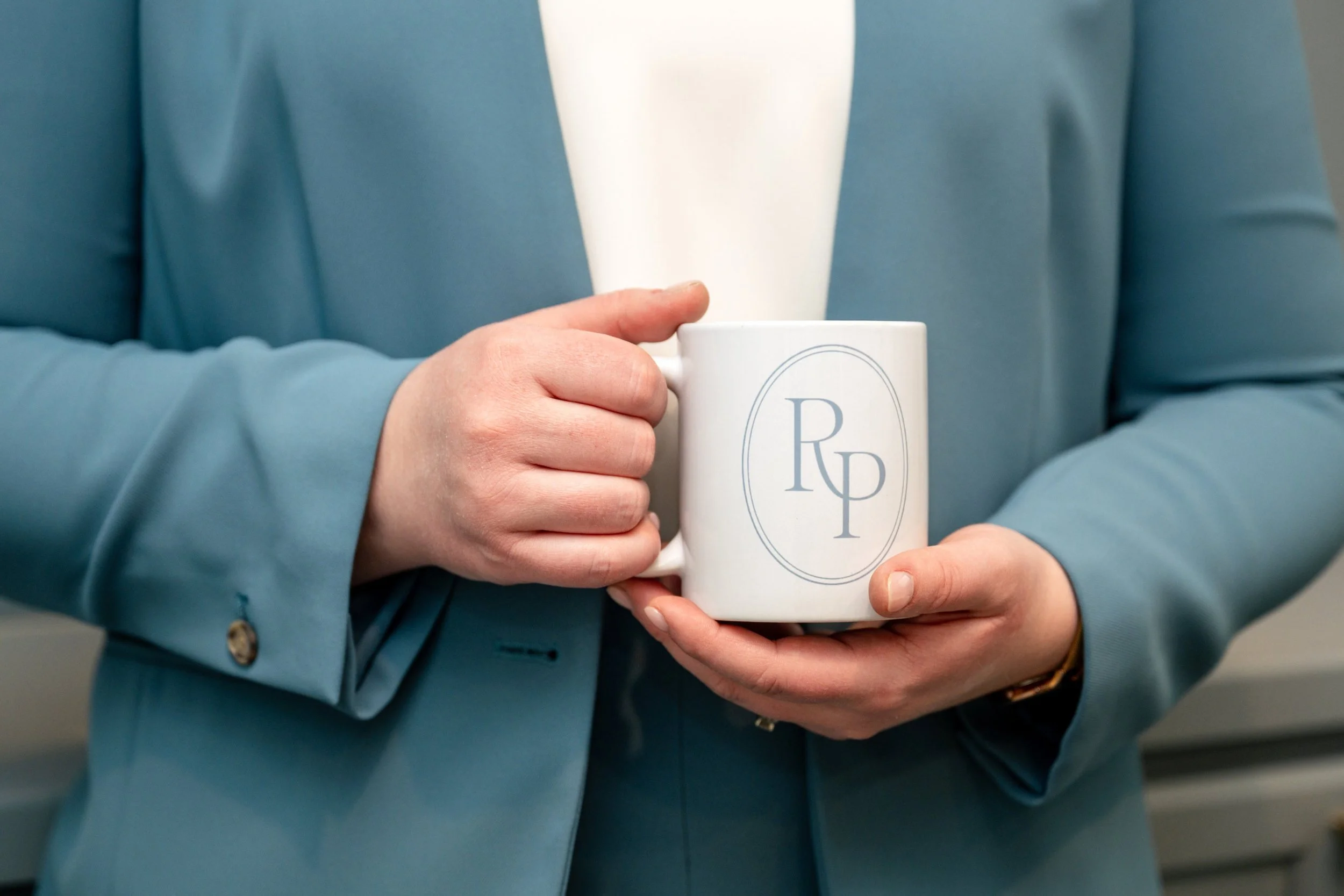 Person holding a white ceramic mug with a logo featuring the letters 'R' and 'P' inside a circle, dressed in a light blue blazer and a white shirt, in an indoor setting.