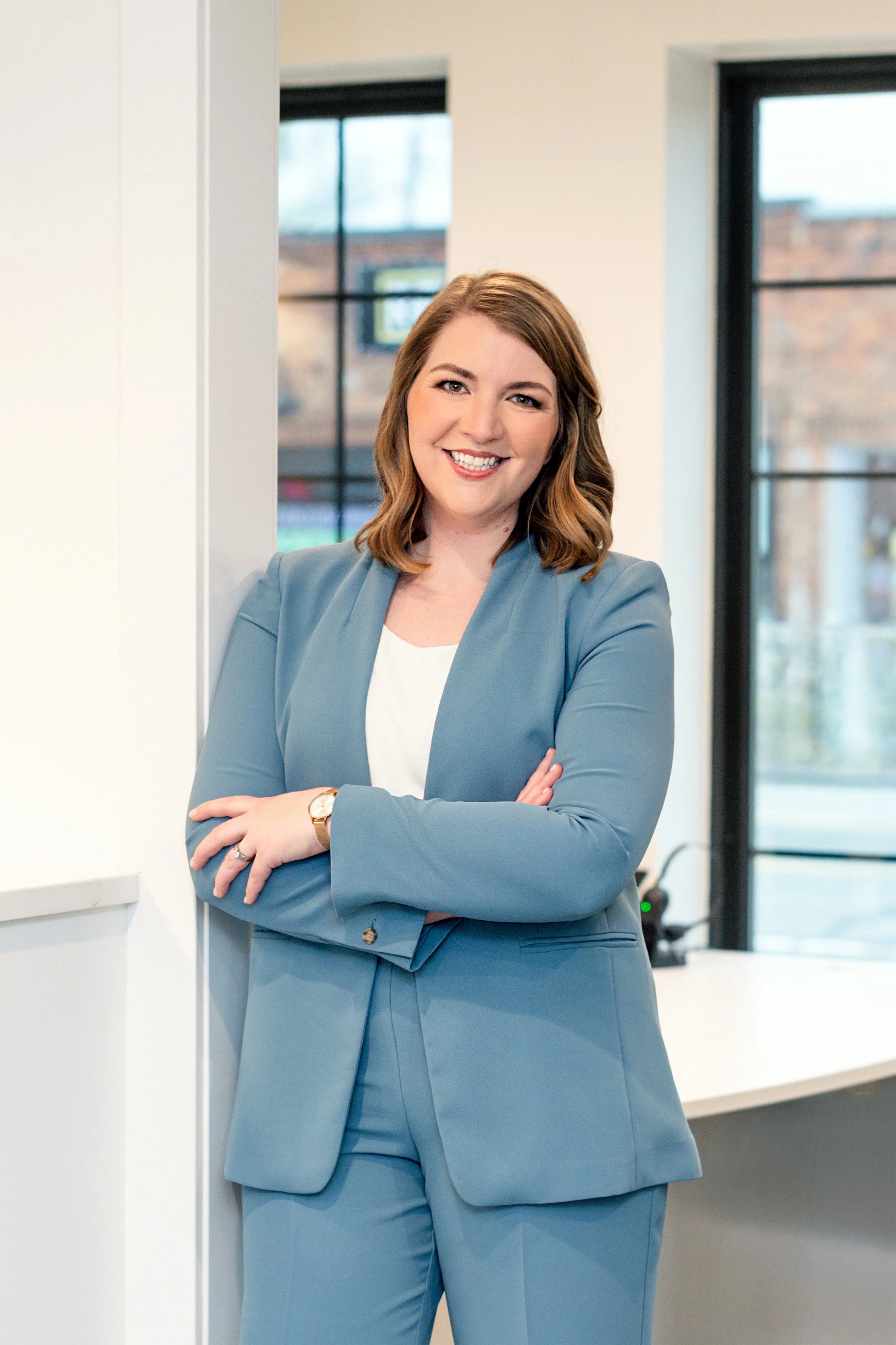 A woman with shoulder-length wavy hair, dressed in a blue suit, standing in a modern office with large windows, smiling with arms crossed.