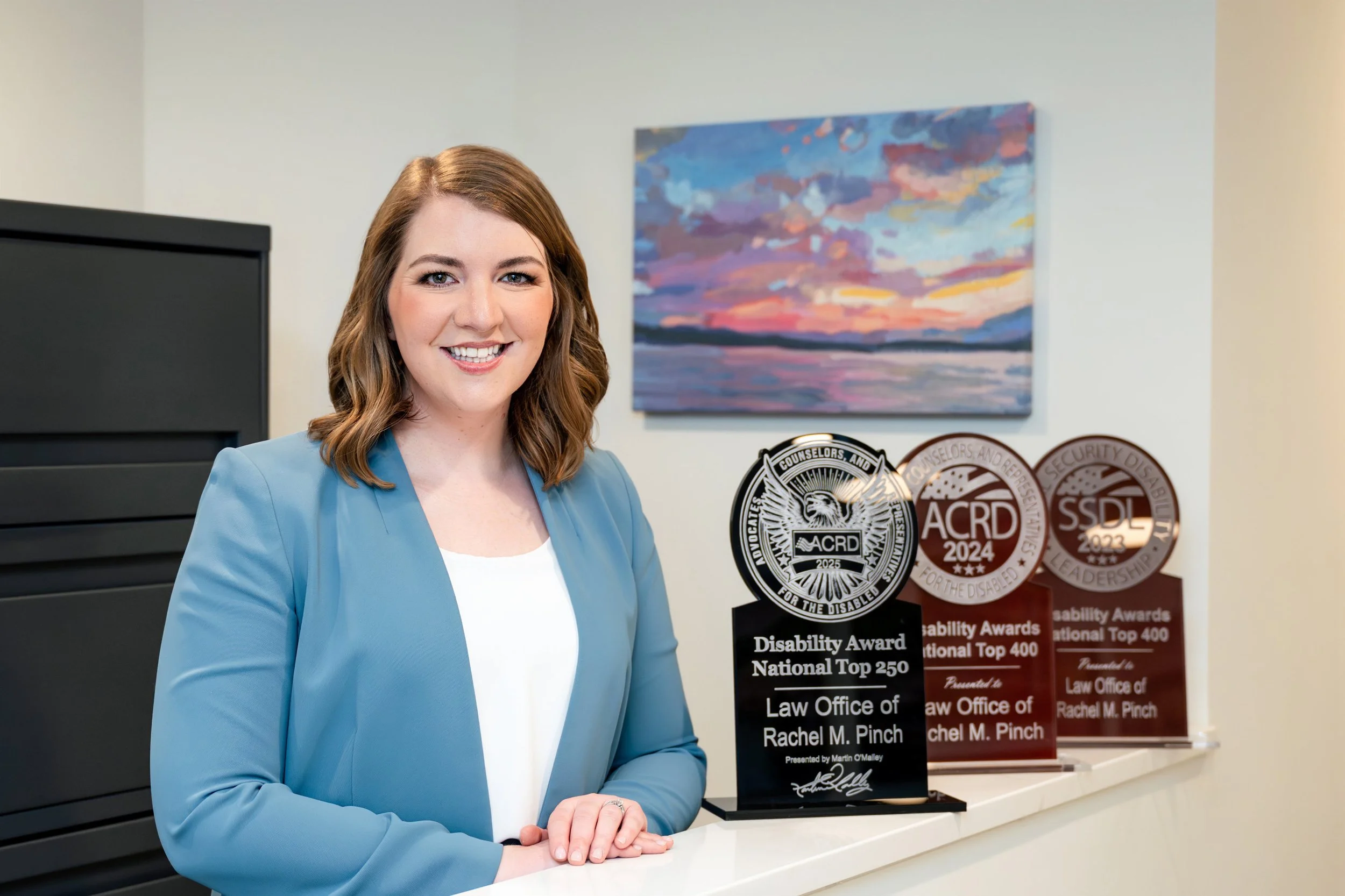 A woman with shoulder-length light brown hair wearing a light blue blazer smiling at the camera, standing in front of award plaques and a colorful landscape painting.