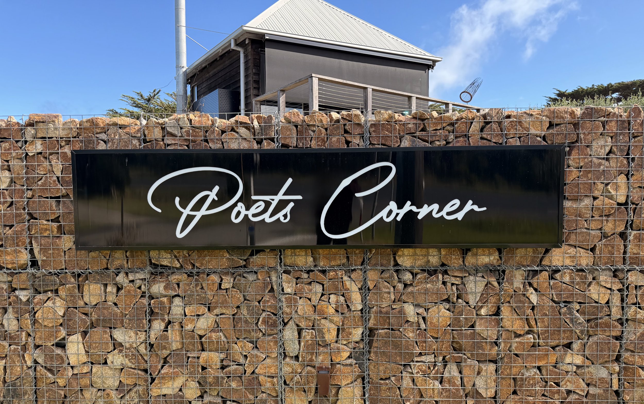 A black sign with white cursive text reading 'Poets Corner' is mounted on a wire mesh fence filled with rocks. Behind the fence, there is a building with a metal roof and wood siding, and a blue sky with some clouds.