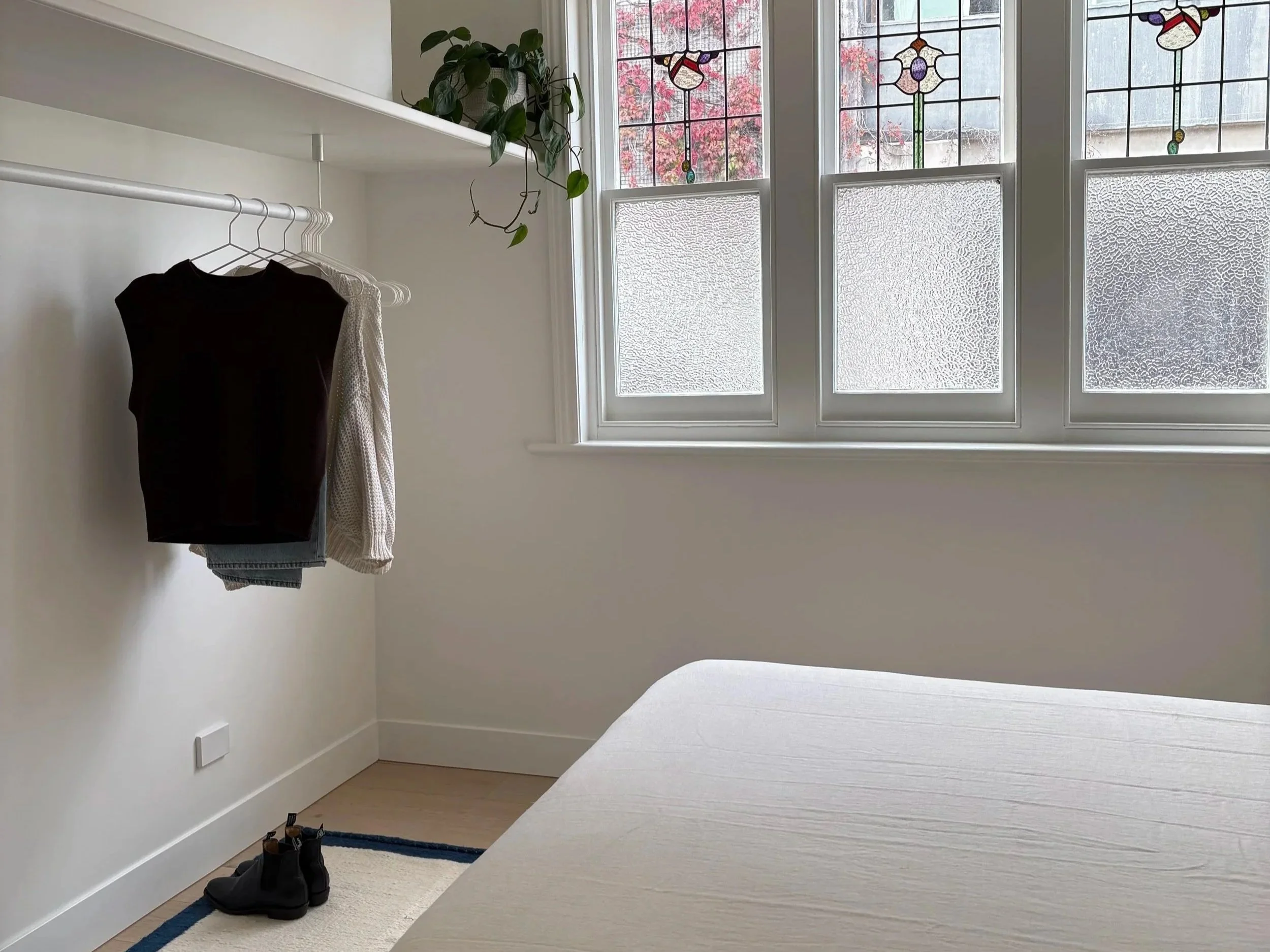 A corner of a room with three tall stained glass windows showing a tree with red leaves outside. Inside, a shelf holds a potted plant, and a clothing rack with black, white, and patterned clothing. A pair of black shoes is on the floor near the wall,