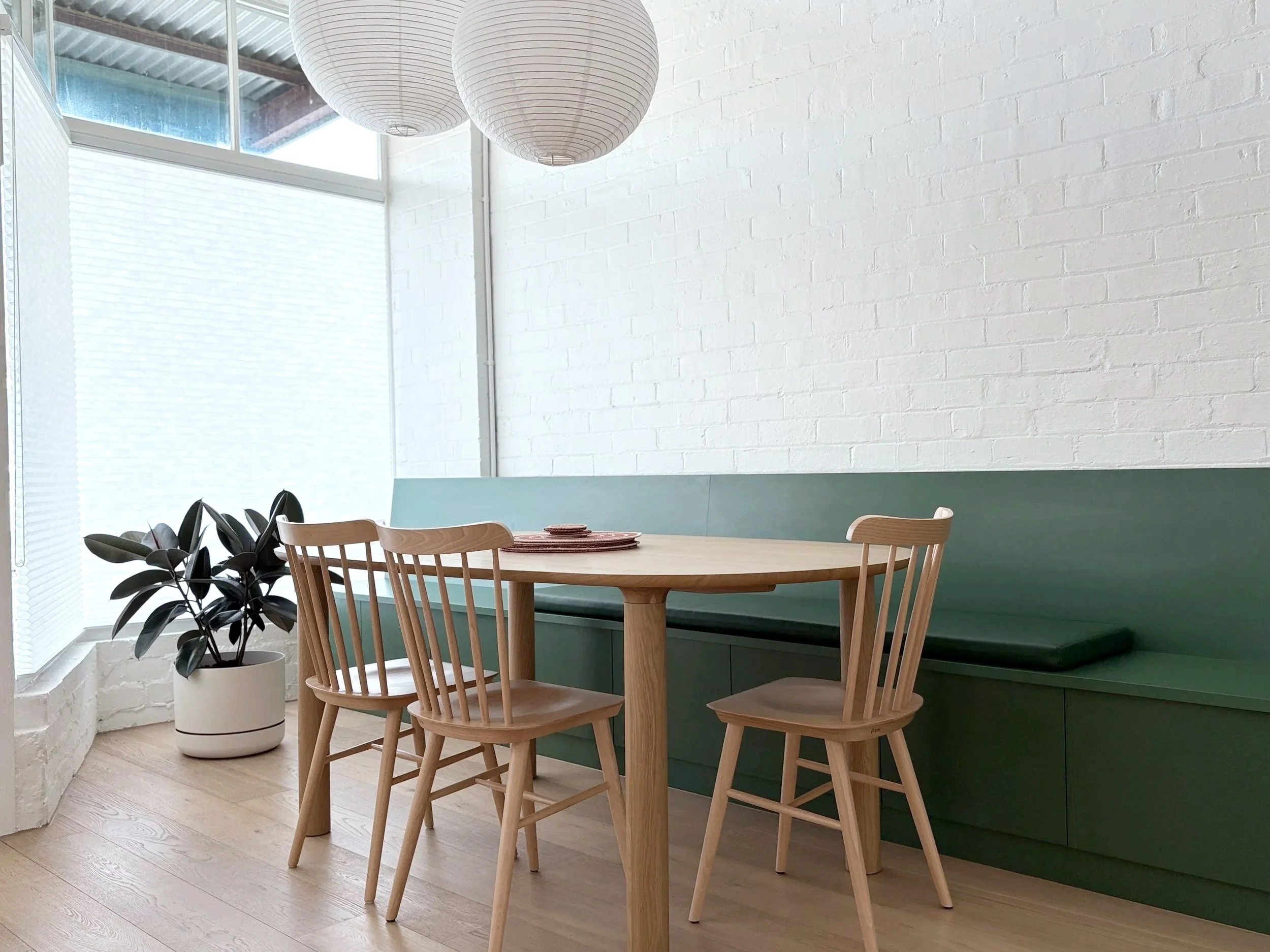 A dining room area with a round wooden table and four matching wooden chairs. A potted plant sits near a large window with blinds, and two white paper lantern pendant lights hang from the ceiling. The walls are painted white with a textured brick pat