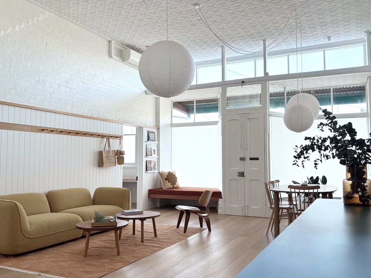Bright living room with white brick walls, beige sofa, wooden coffee tables, round hanging paper lantern lights, a corner bench with cushion, dining table with chairs, large windows, and potted plant on a sideboard.