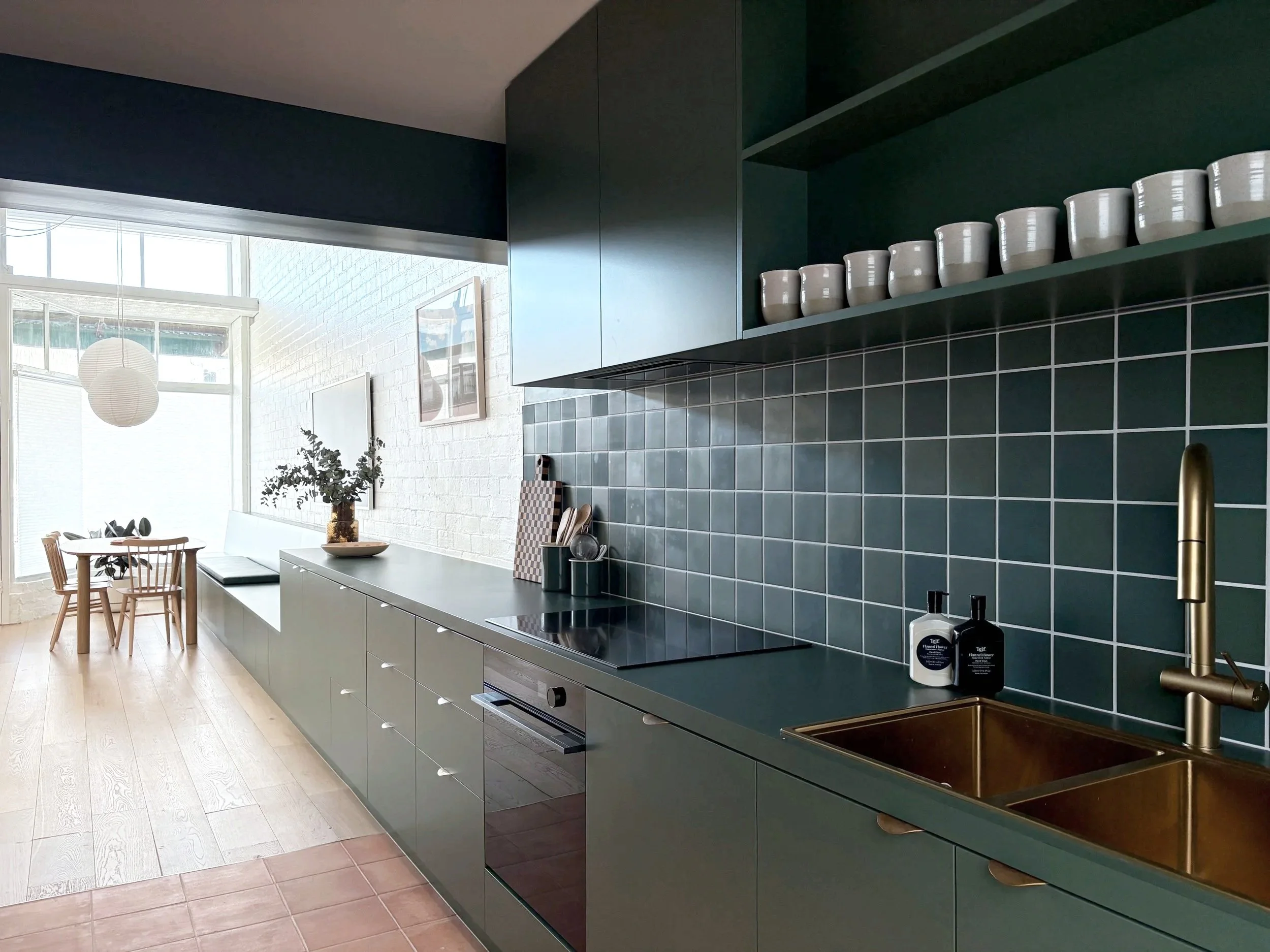 Modern kitchen with dark green cabinets and backsplash, white dishware on open shelf, and light wood flooring with a dining area in the background.