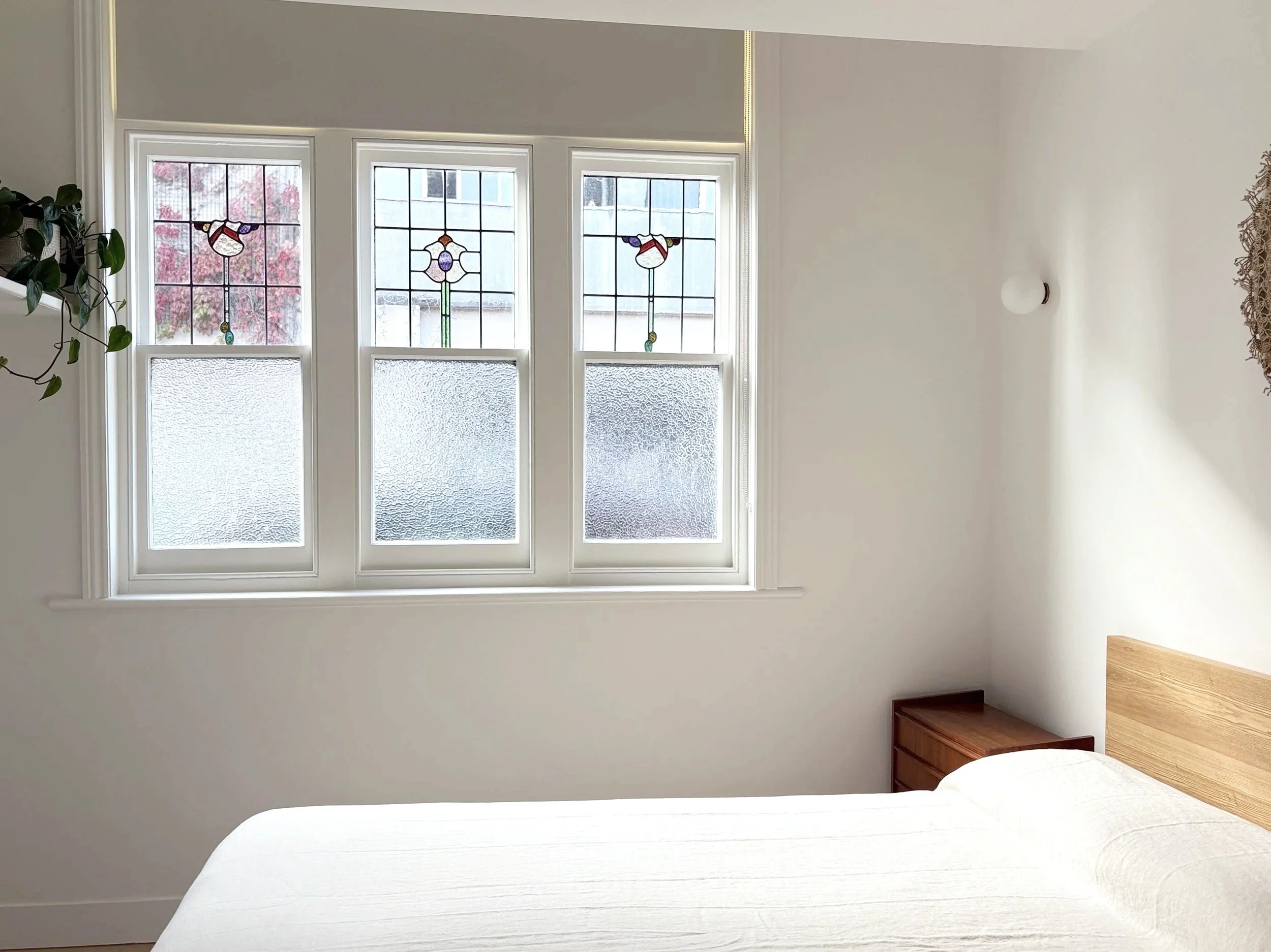 Interior view of a room with three large windows featuring stained glass designs, white walls, a white bed with a wooden headboard, a small wooden nightstand, and a ceiling light fixture.