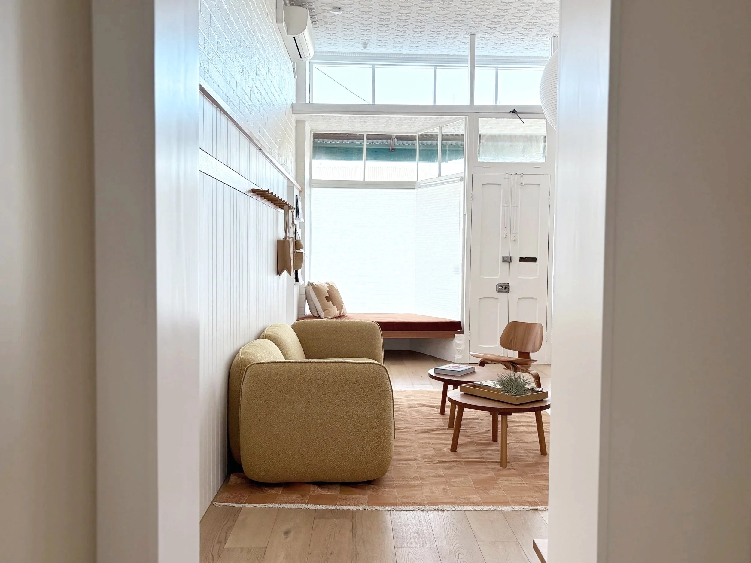 A cozy living room with a beige sofa, a small wooden coffee table with a tray and plant, and a bench beneath a window with white walls and natural light.