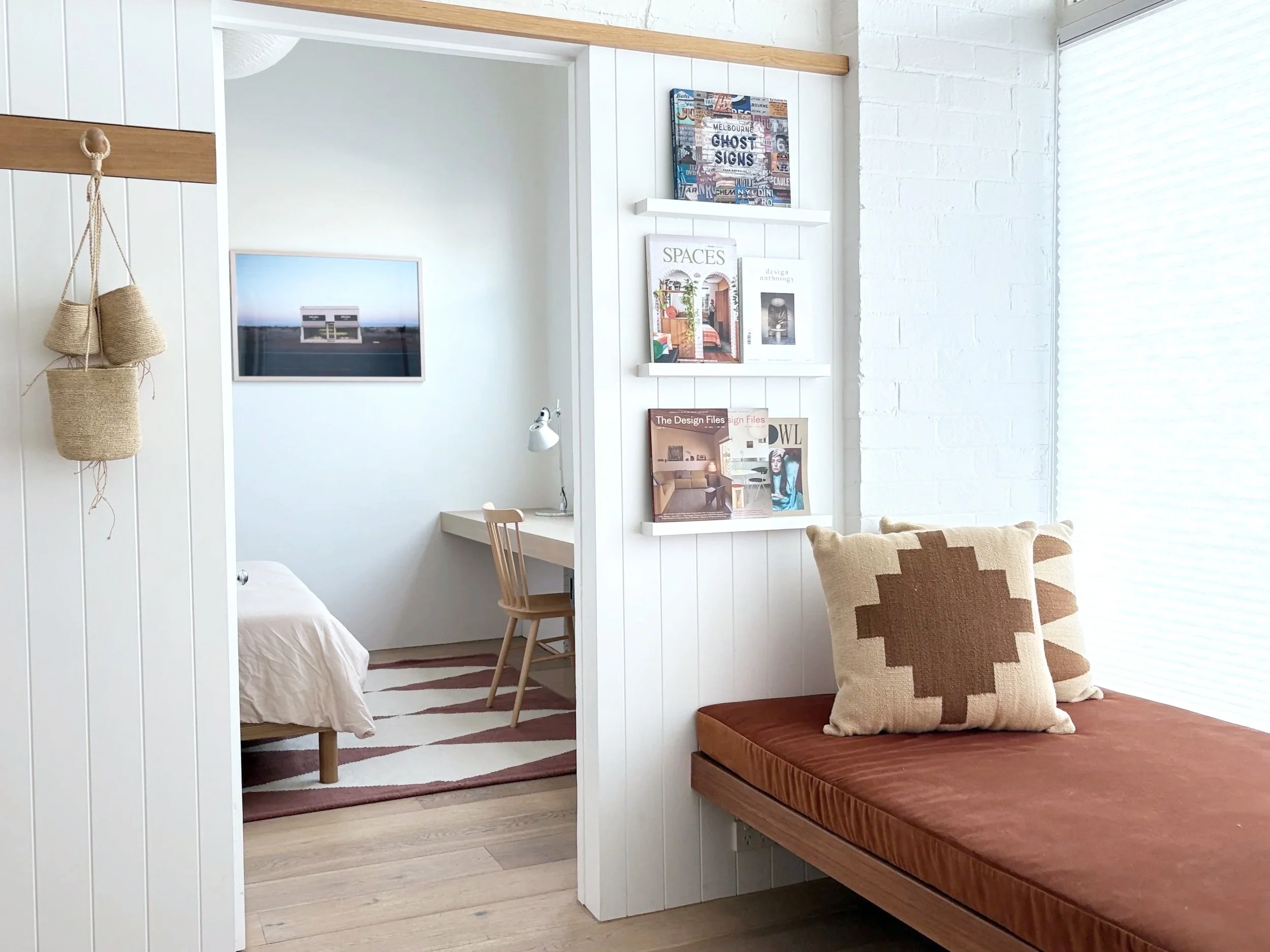 Interior of a modern small apartment with a bedroom visible through an open doorway. The bedroom has a bed with white bedding, a wall-mounted picture, and a window with a view. In the living area, there is a cushioned bench with two decorative pillow