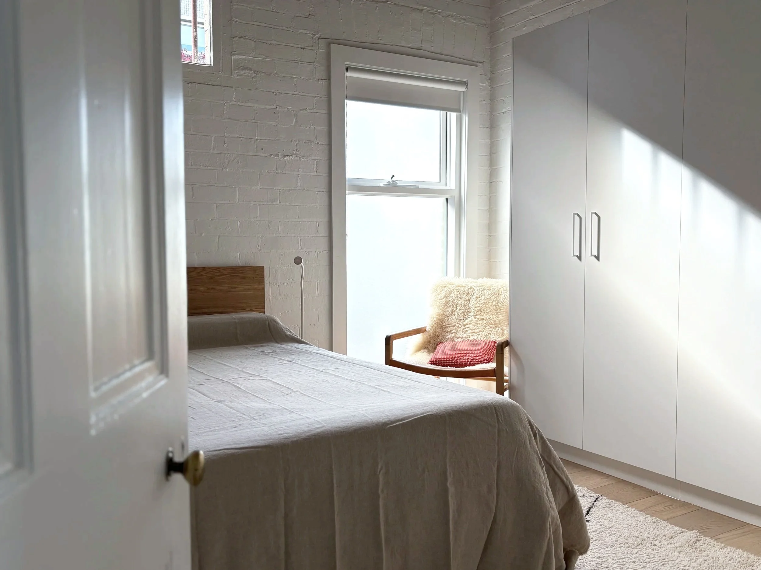 View into a bedroom through a partially open door, showing a bed with a beige bedspread, a white brick wall, a window with frosted glass, a white wardrobe, and a chair with a red cushion and a beige fluffy throw.