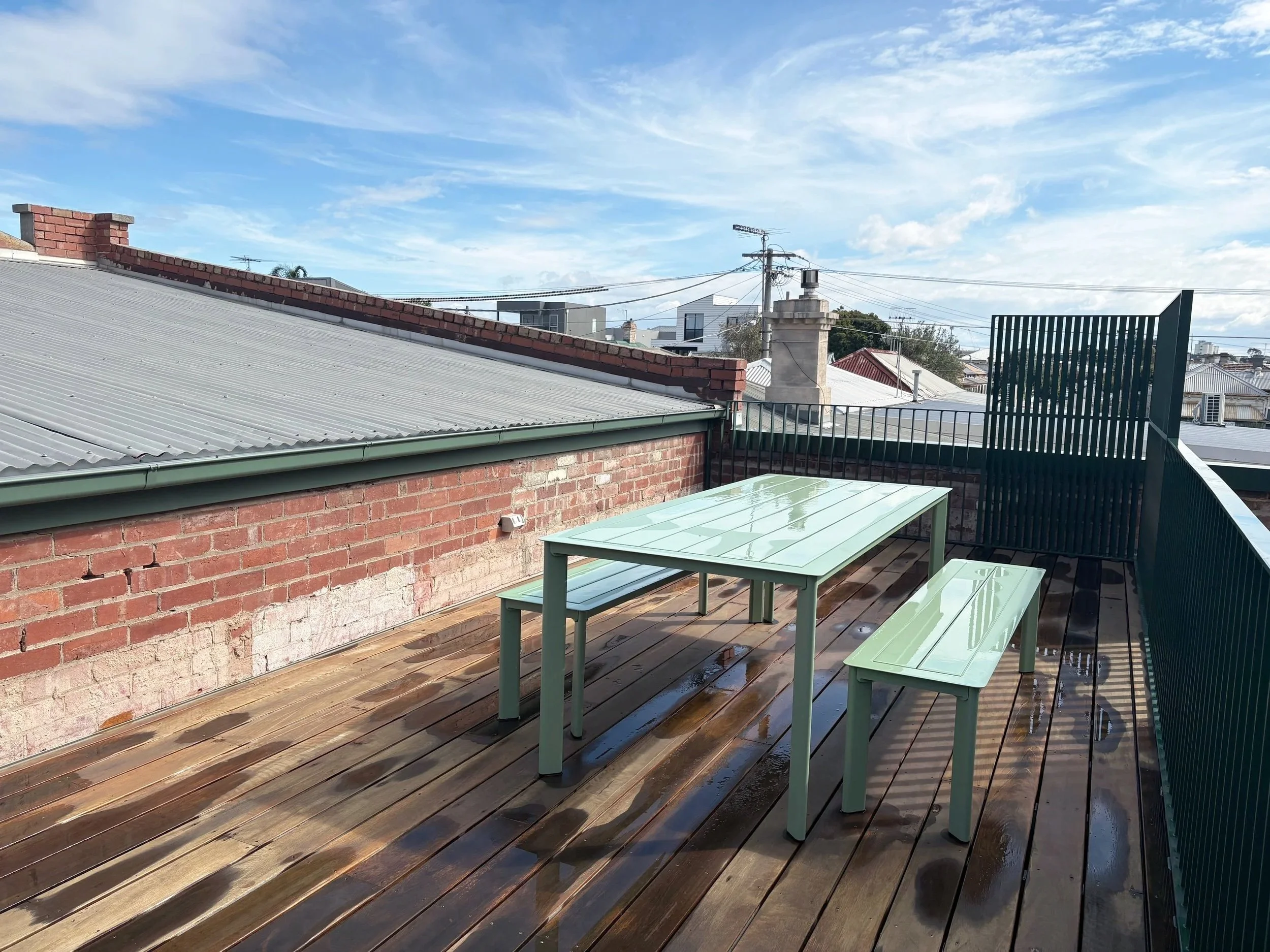 Rooftop terrace with wooden deck and green tables, brick walls, metal fencing, and neighboring rooftops with sky and clouds overhead.