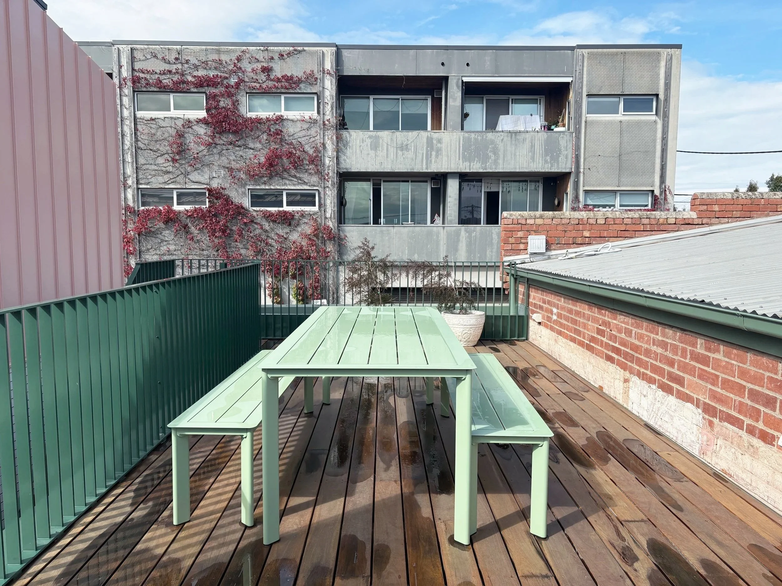 An outdoor balcony with green dining table and benches, wooden deck flooring, metal railing, adjacent brick and metal buildings, and a partly cloudy sky in the background.