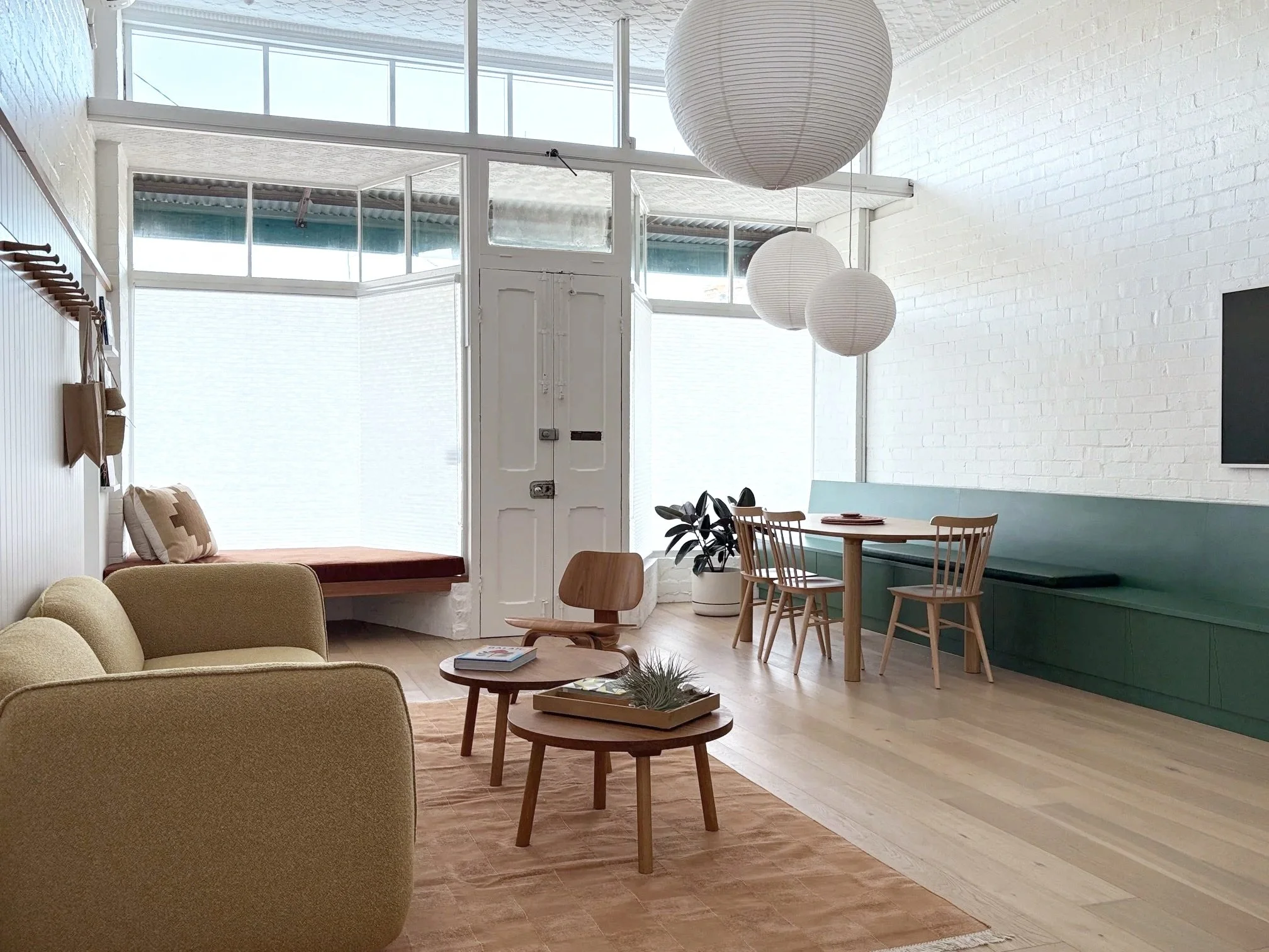 Interior of a modern, minimalist living room with light-colored wooden flooring, beige upholstered chairs, a round wooden coffee table, and a built-in seating area with cushions beneath large windows allowing natural light. The walls are white brick,