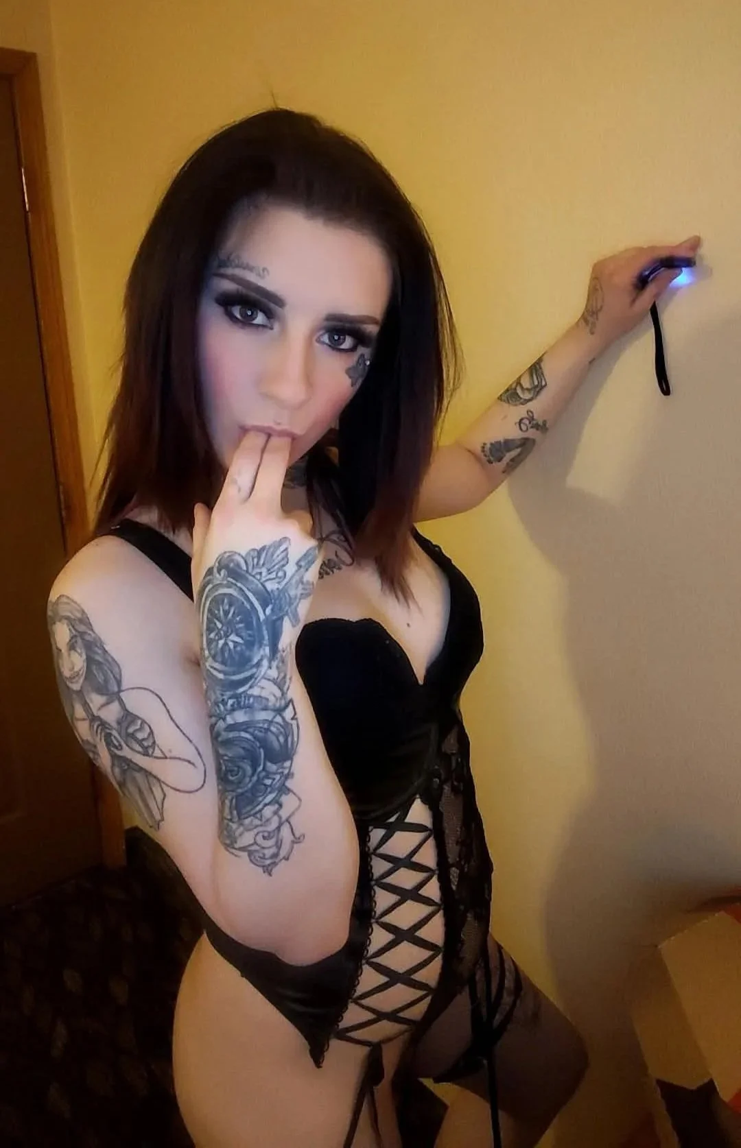 A woman with dark hair and tattoos on her arms and torso wearing black lace lingerie, posing indoors against a yellow wall.